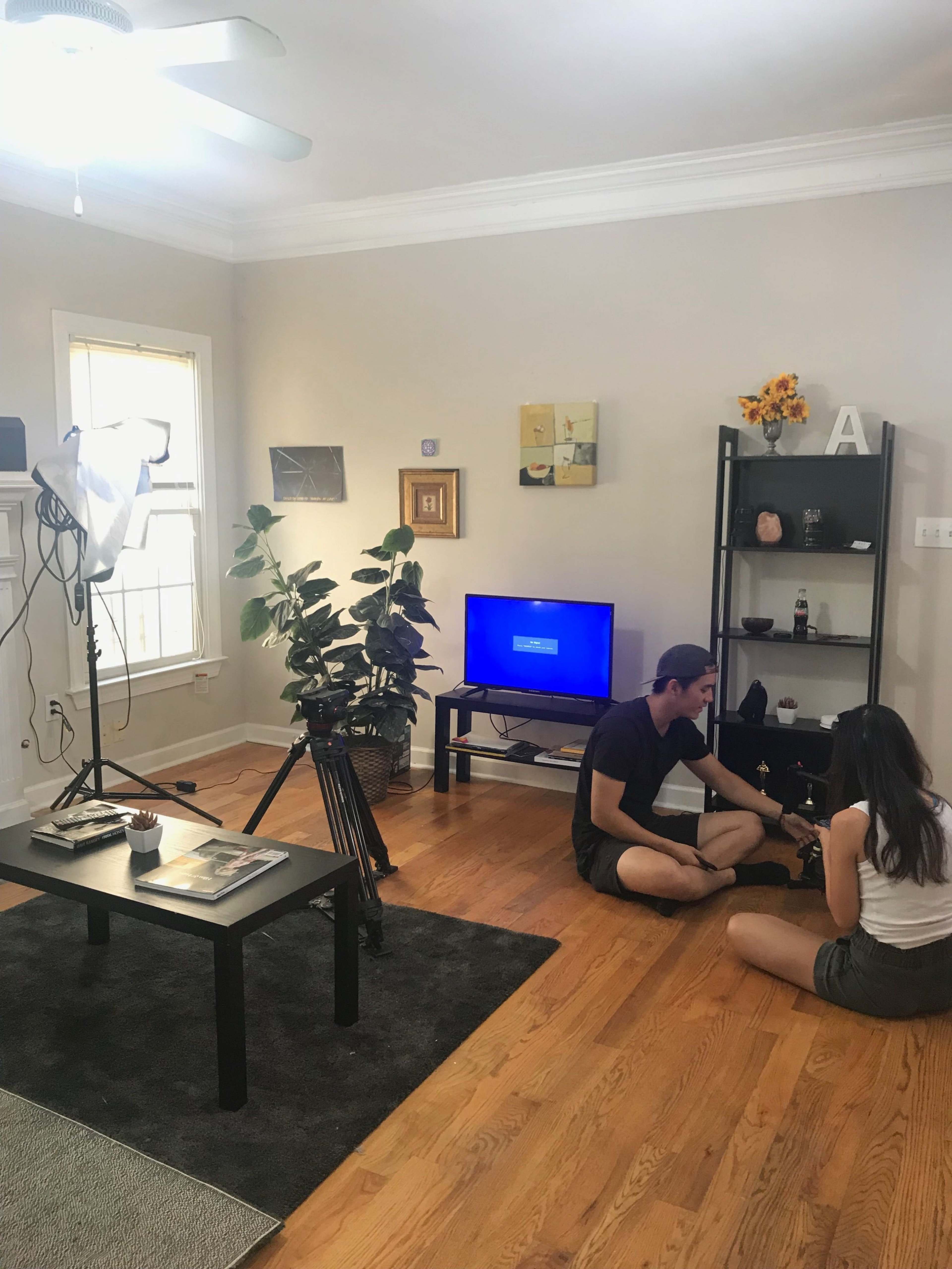 Two people are setting up equipment in a well-lit living room with a television and a bookshelf.
