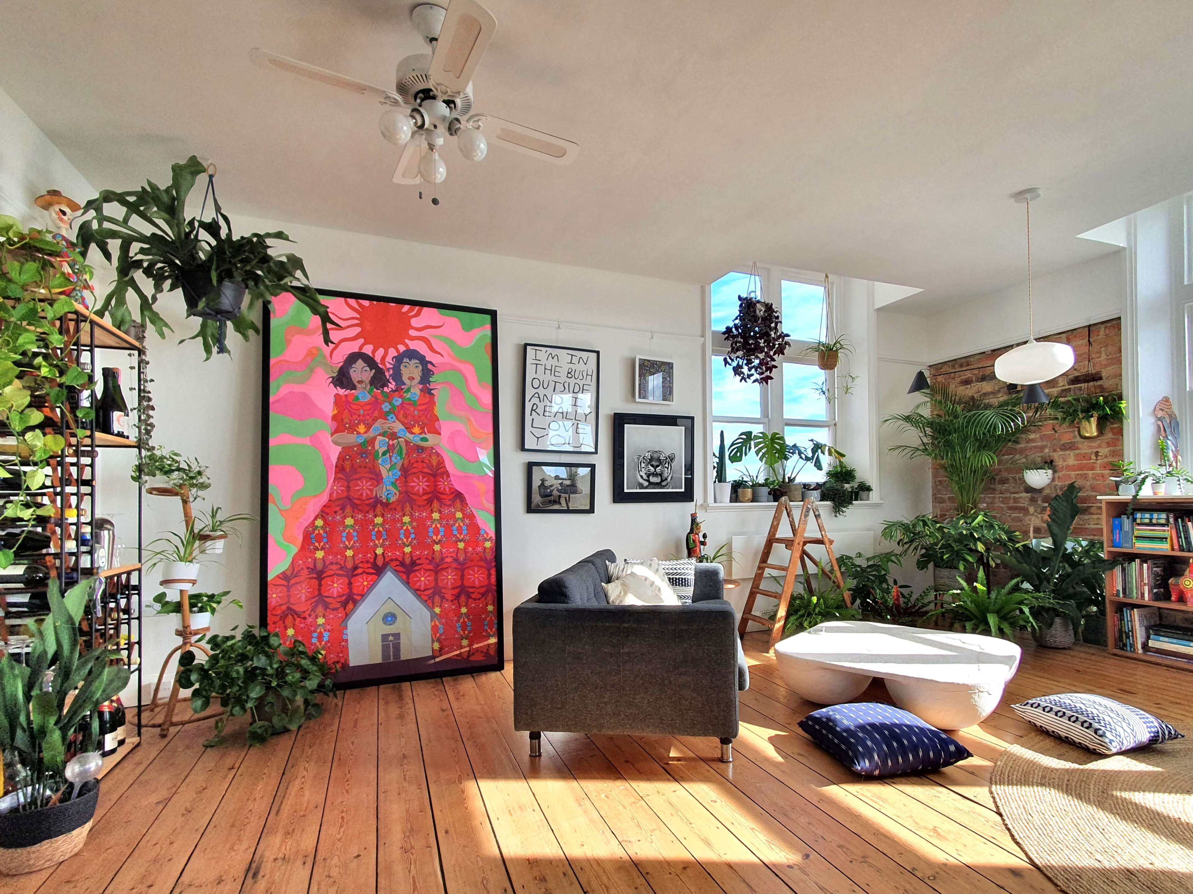 A bright, modern living room with large windows, featuring a colorful mural, houseplants, and a mix of furniture including a sofa and a coffee table.