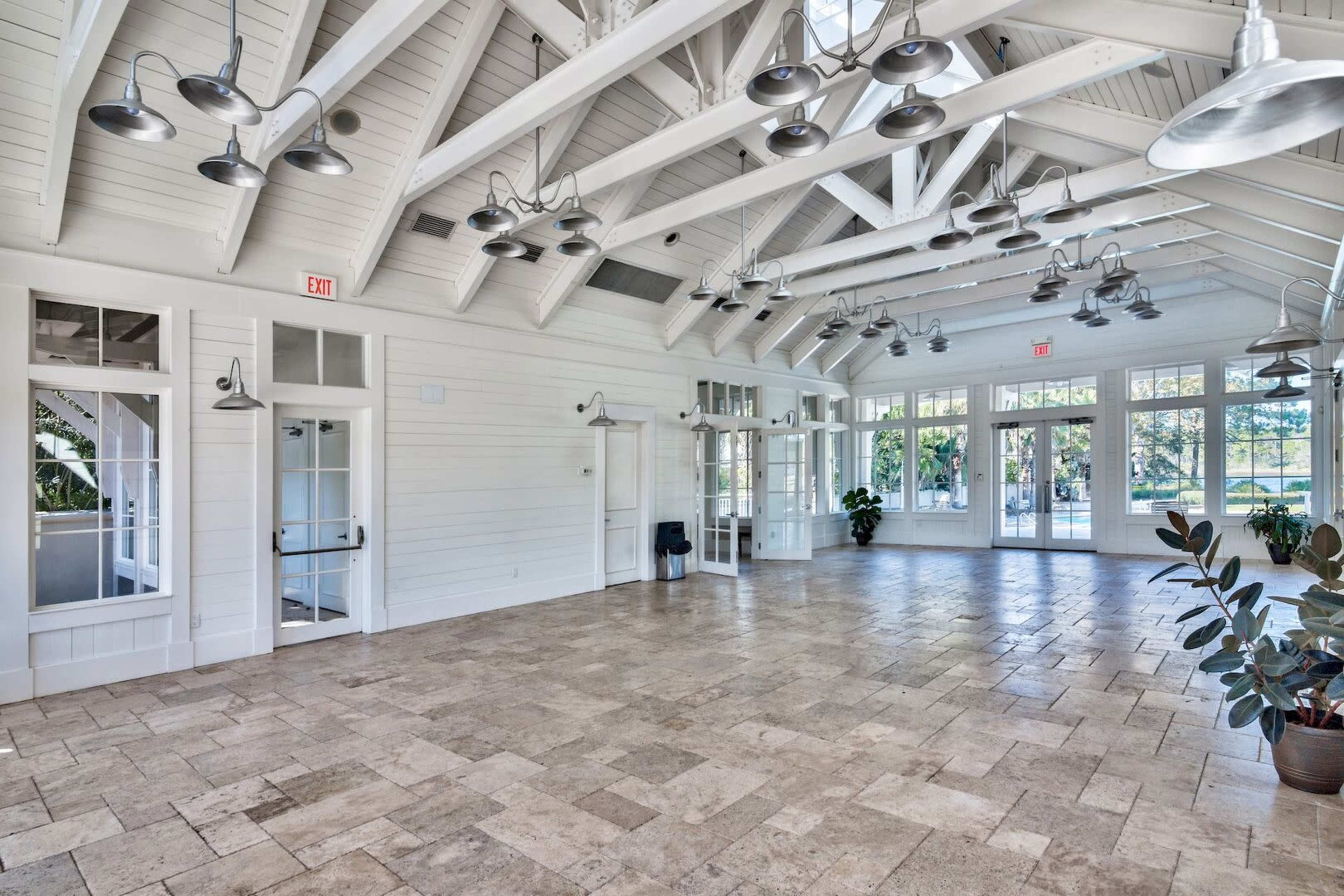 A spacious interior with high, white-beamed ceilings, large windows, and stone flooring, featuring minimal furnishings and greenery.