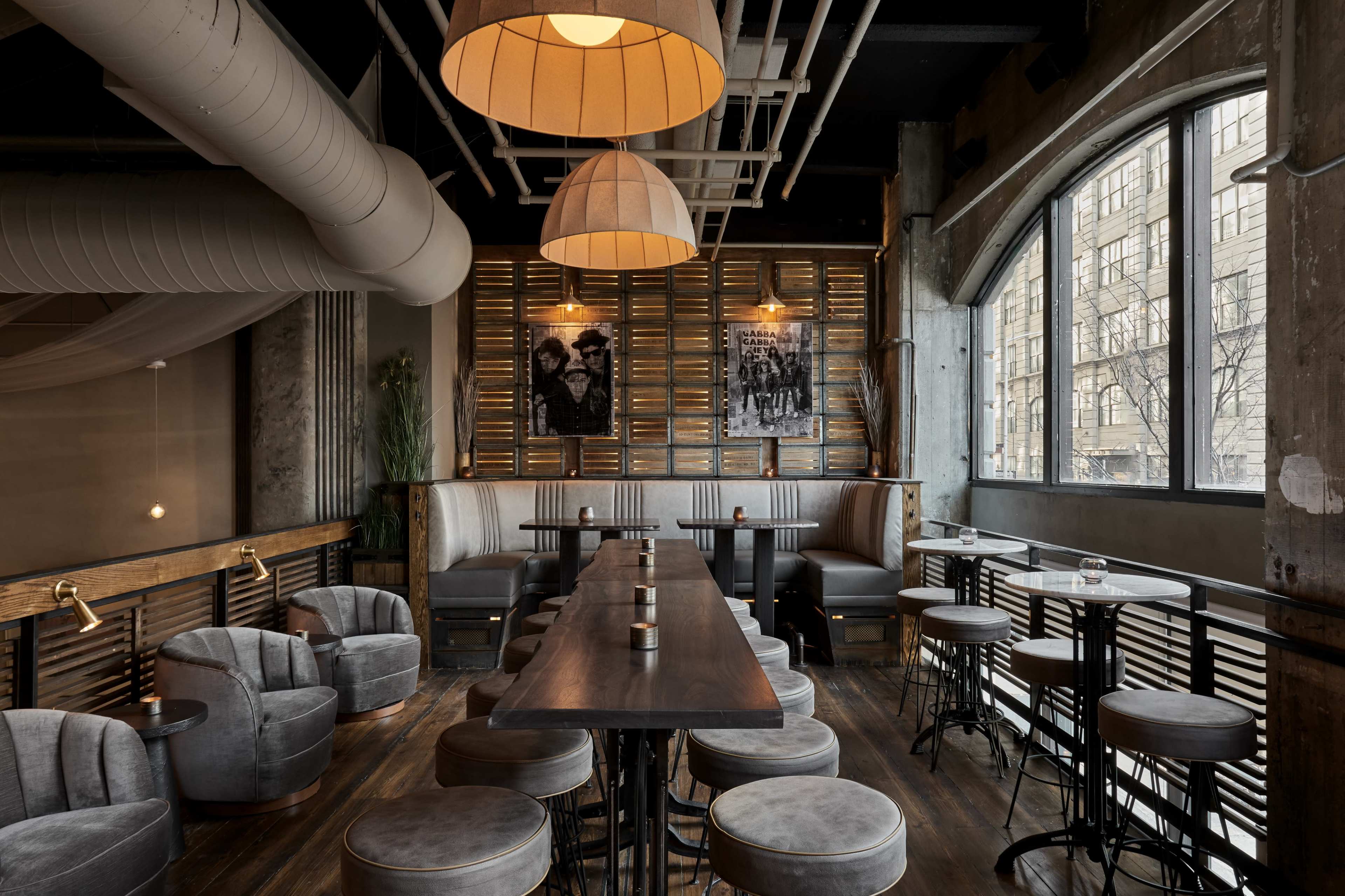 A modern restaurant interior with gray upholstered chairs and a long wooden table, illuminated by pendant lights and featuring industrial design elements.