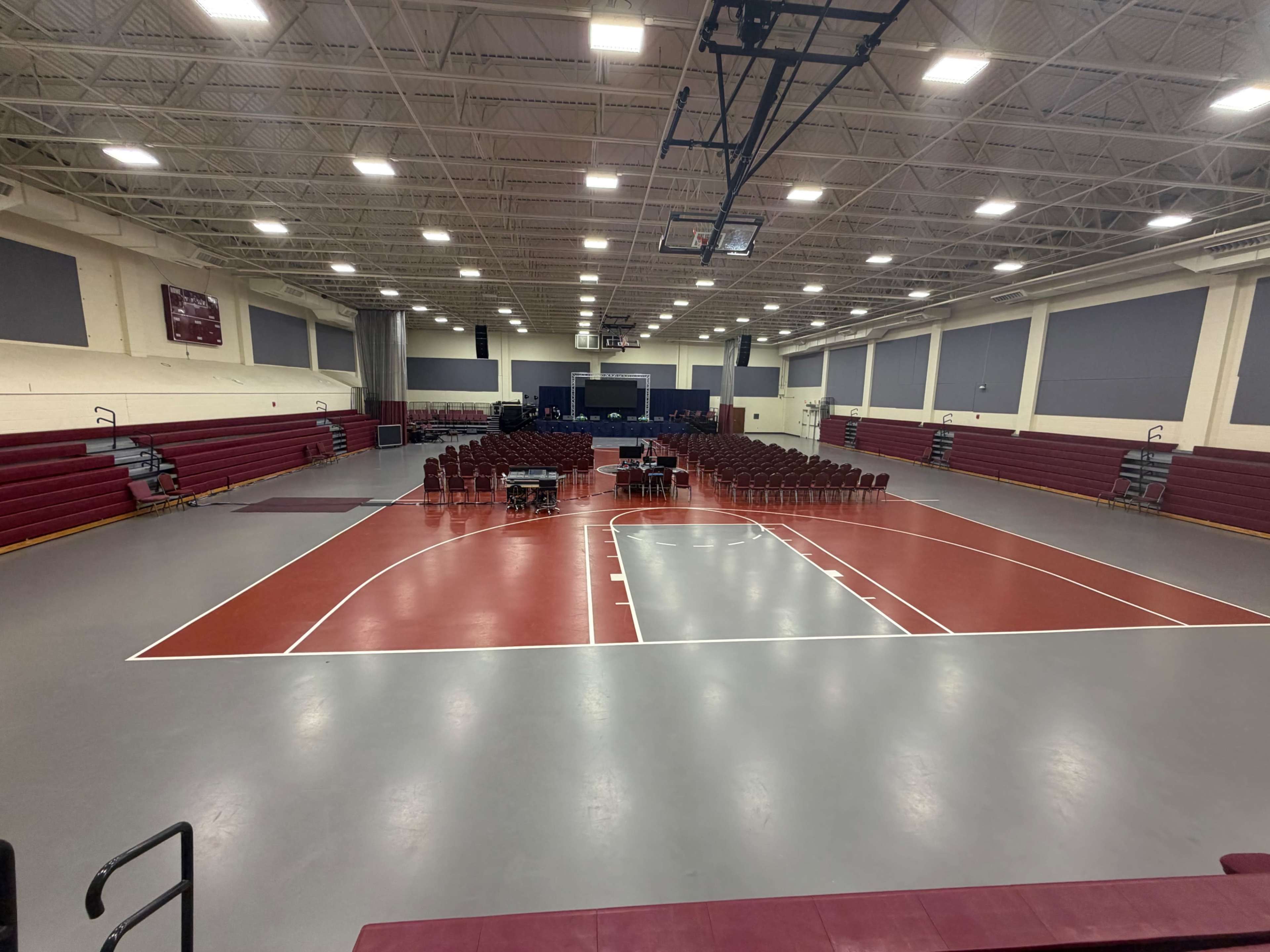 The image shows a large gymnasium with a basketball court and rows of chairs set up for an event.