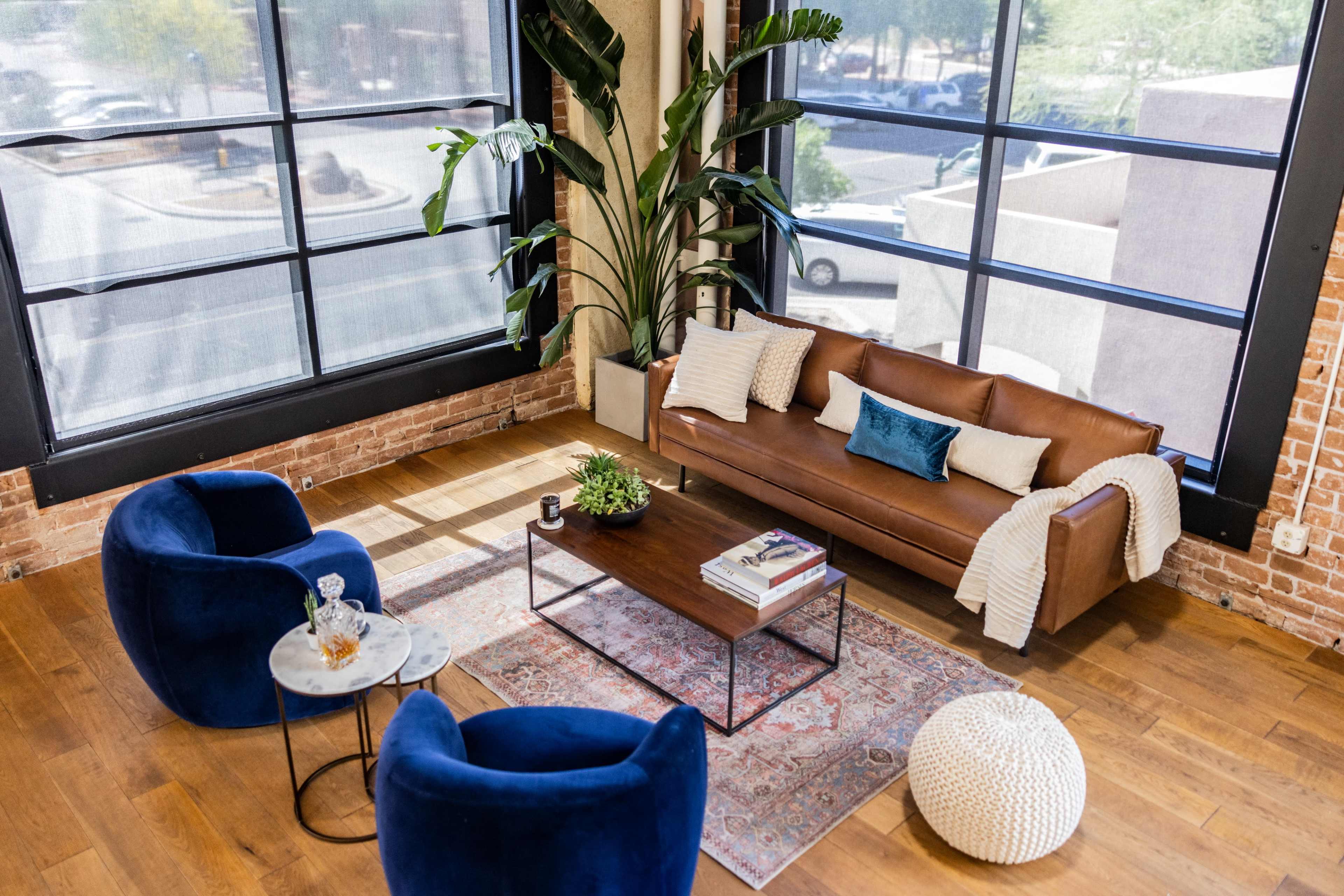 A modern living room features a brown leather sofa, two blue chairs, a coffee table with books, and a large window with lush greenery outside.