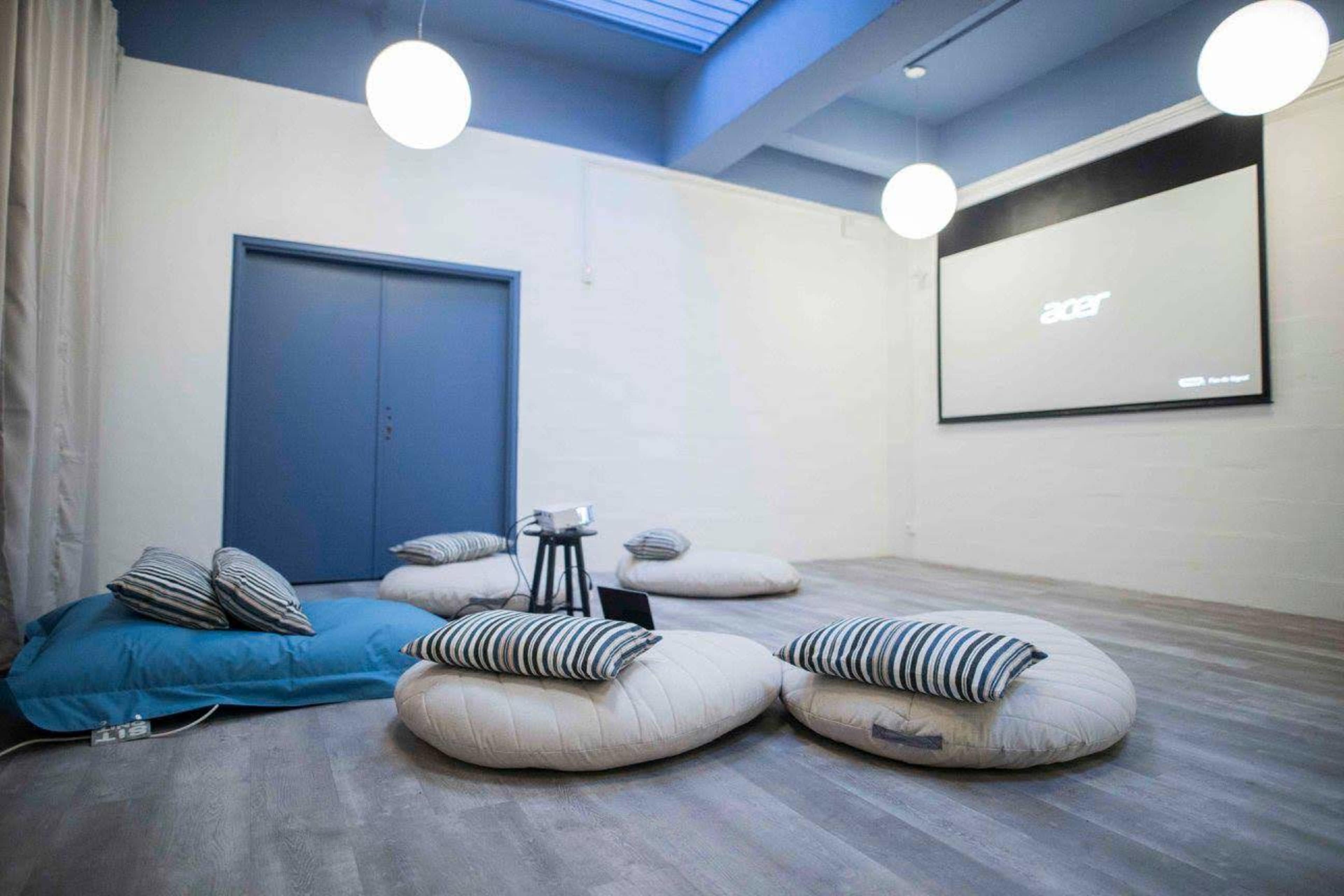 A minimalist room features several large floor cushions, a small table, and a projector screen on a white wall.