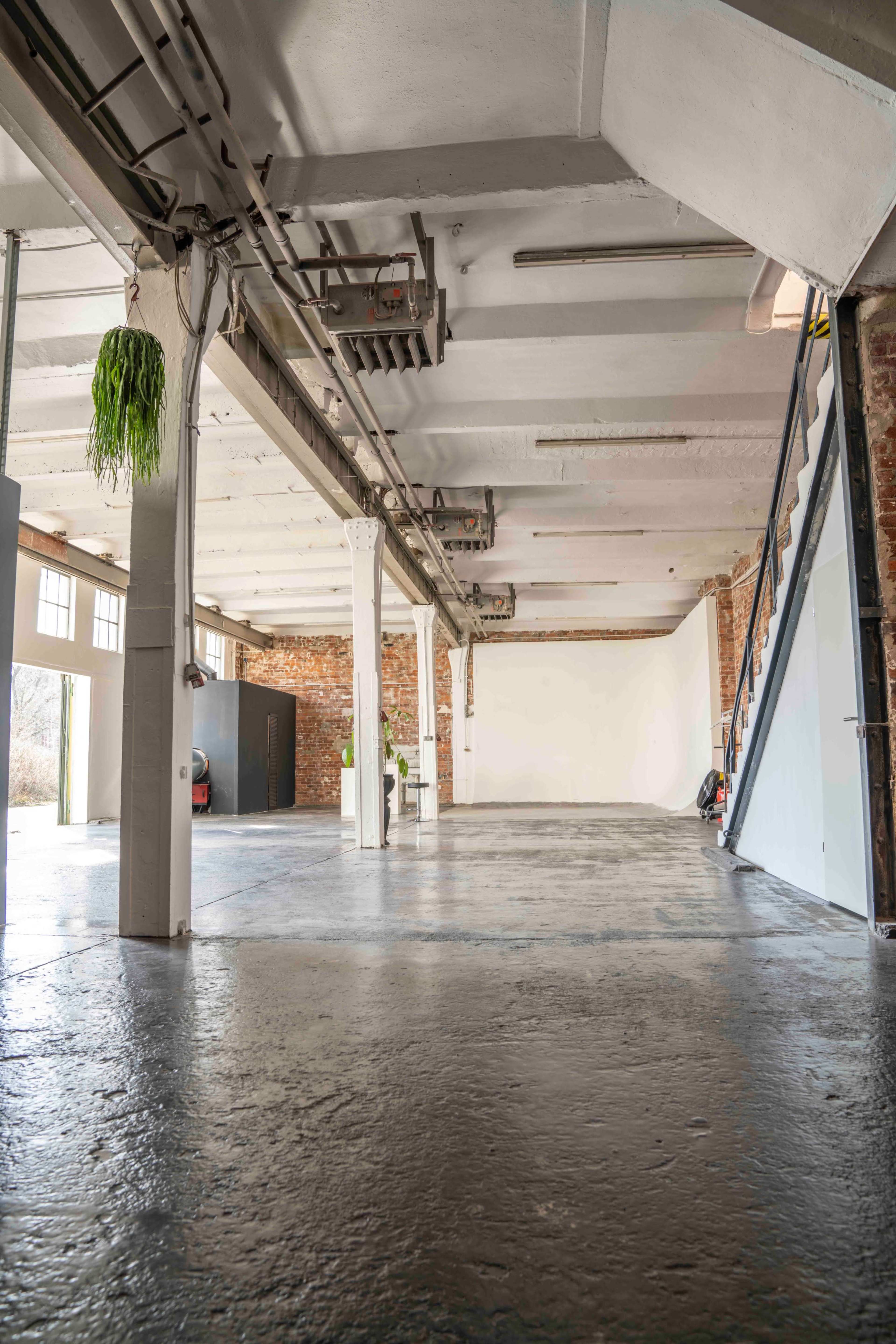 The image shows a spacious industrial-style room with exposed brick walls, polished concrete floor, and an open layout featuring a staircase in the corner.