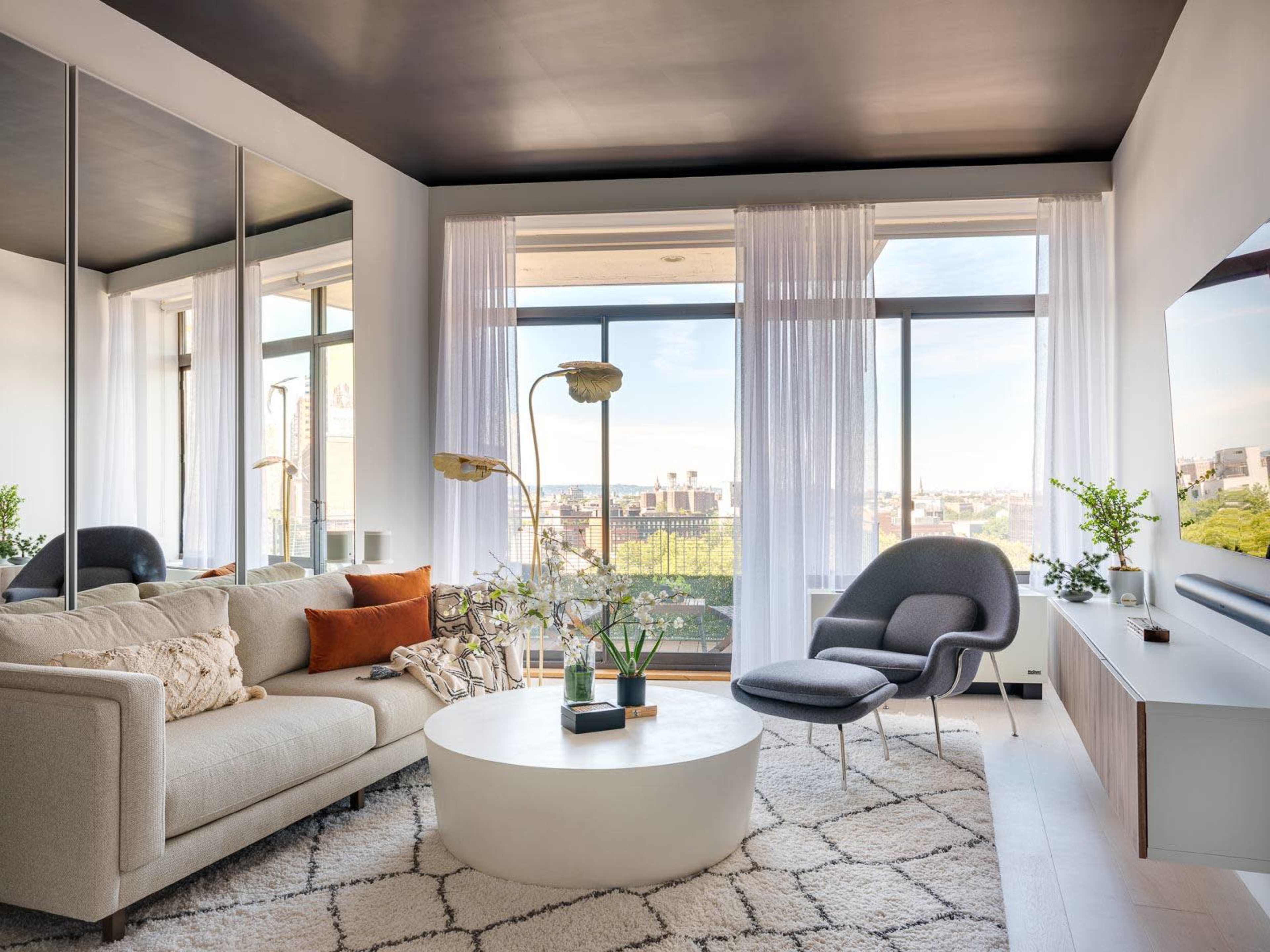 The image shows a modern living room with a large window offering a view, featuring a light-colored sofa, a round coffee table, a gray armchair, and minimalistic decor.