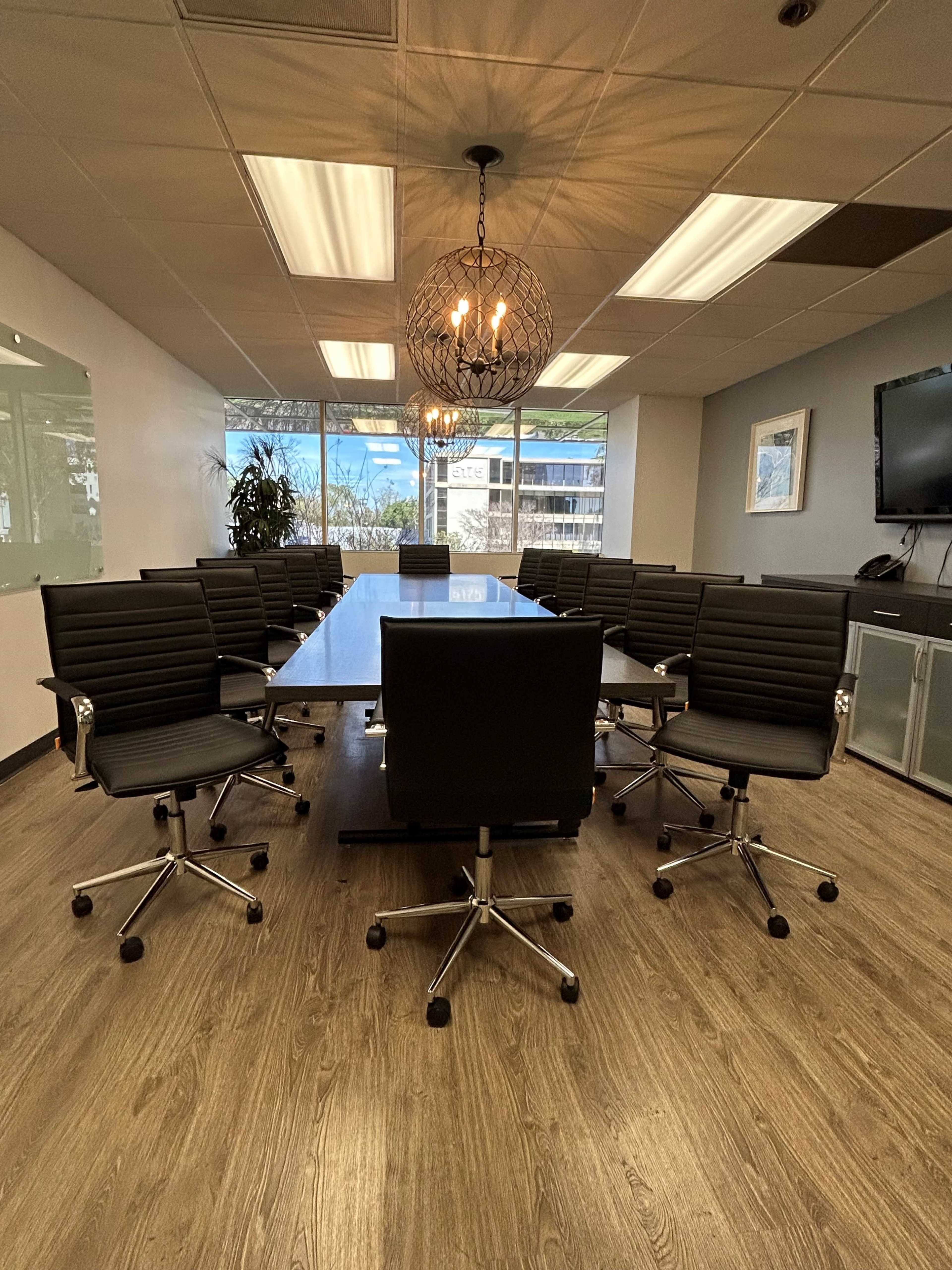 A large conference room features a long glass table surrounded by black swivel chairs, with bright windows and modern lighting overhead.