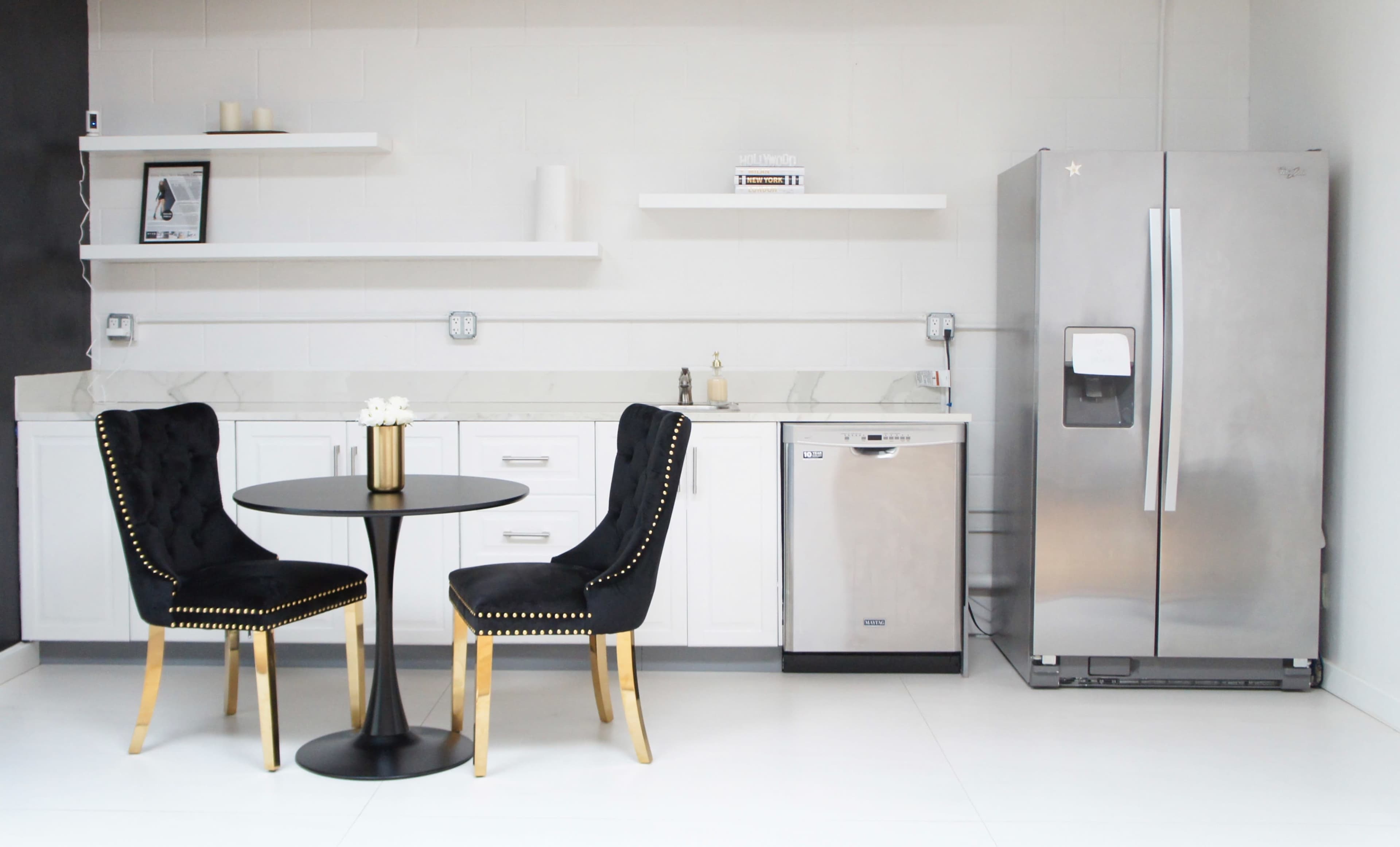 A modern kitchen features a round black table with two upholstered chairs and stainless steel appliances against a light-colored wall.