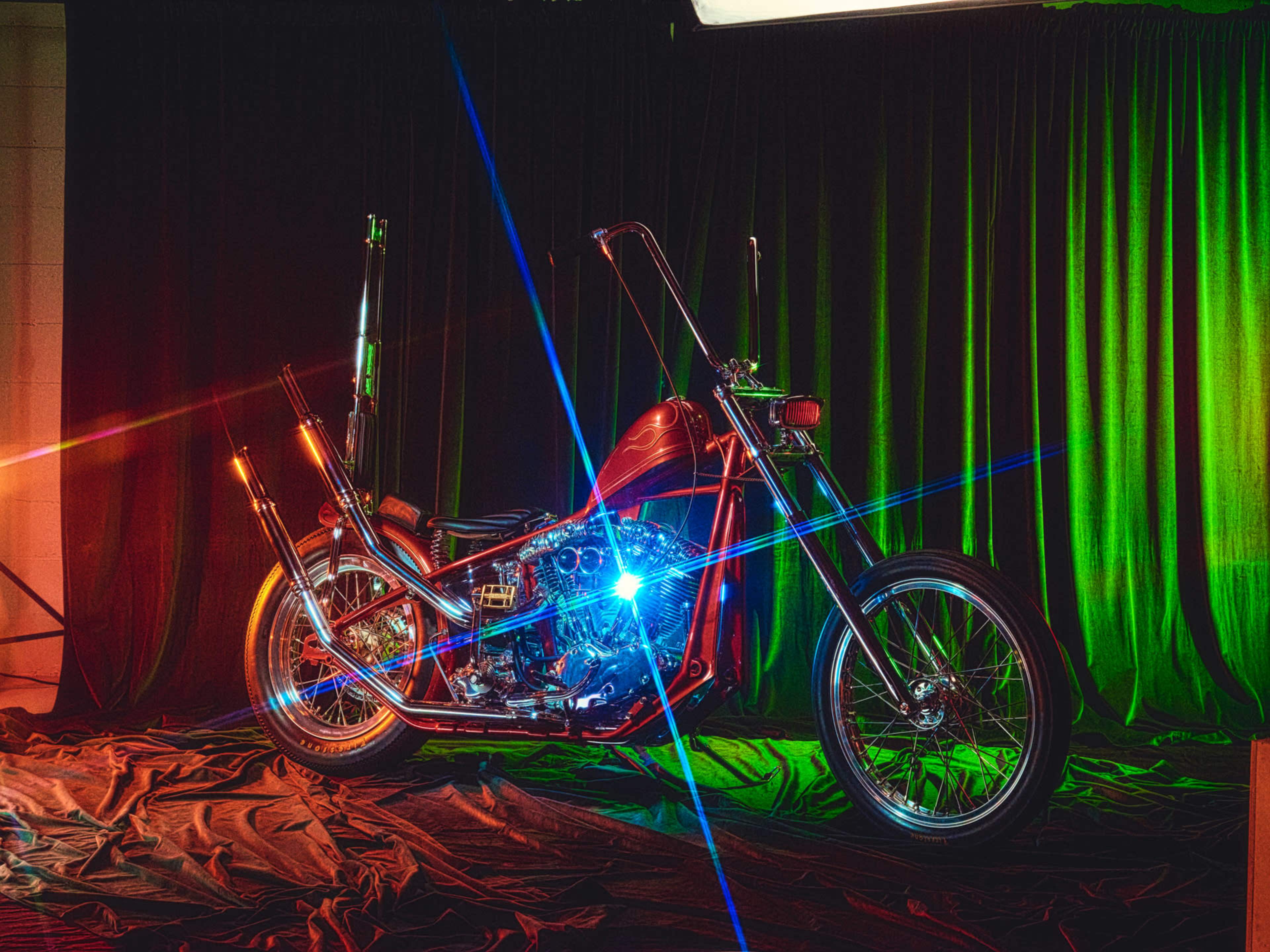 A custom motorcycle is displayed on a textured surface with colorful lighting and a dark curtain backdrop.