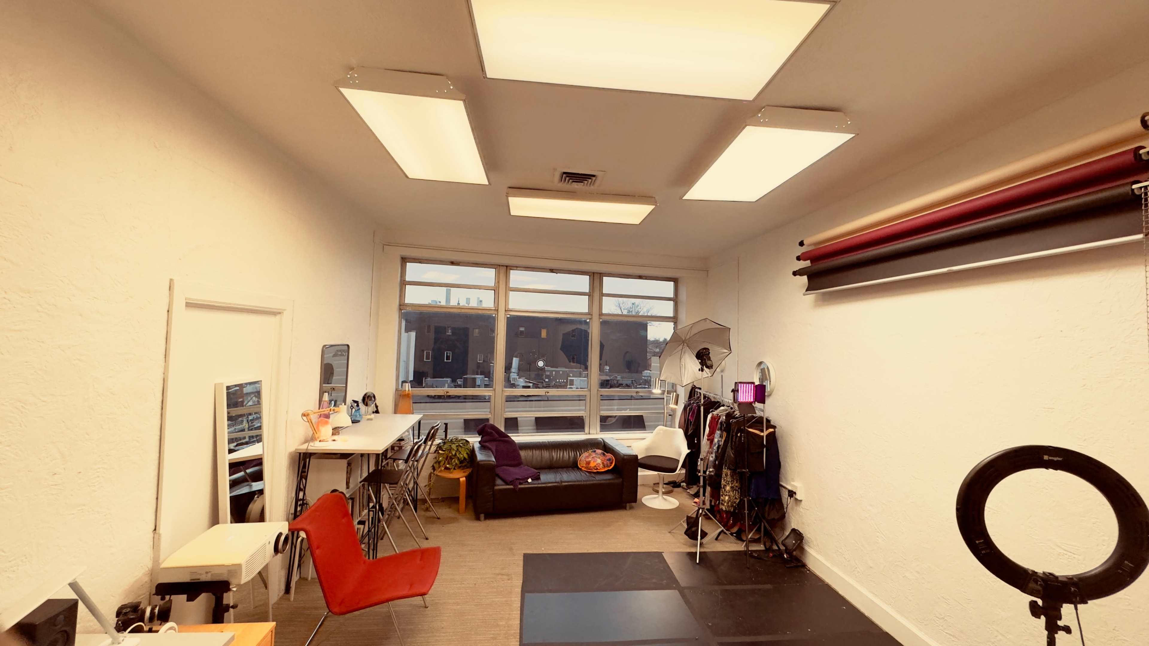 The image shows a brightly lit studio space featuring a seating area, several chairs, a table, and various photography equipment along one wall.