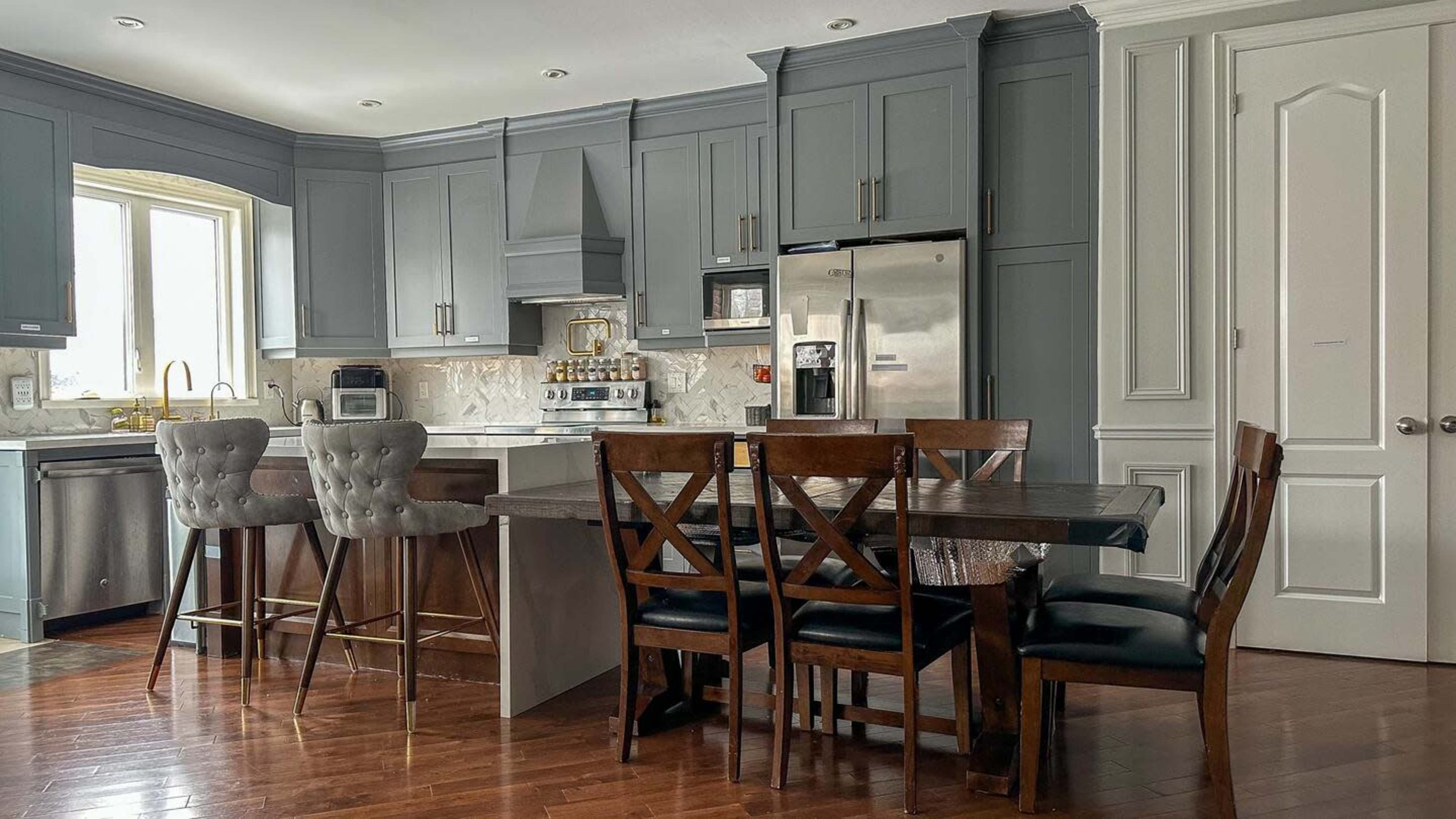 A modern kitchen with gray cabinets, a stainless steel refrigerator, a marble backsplash, and a wooden dining table surrounded by chairs.