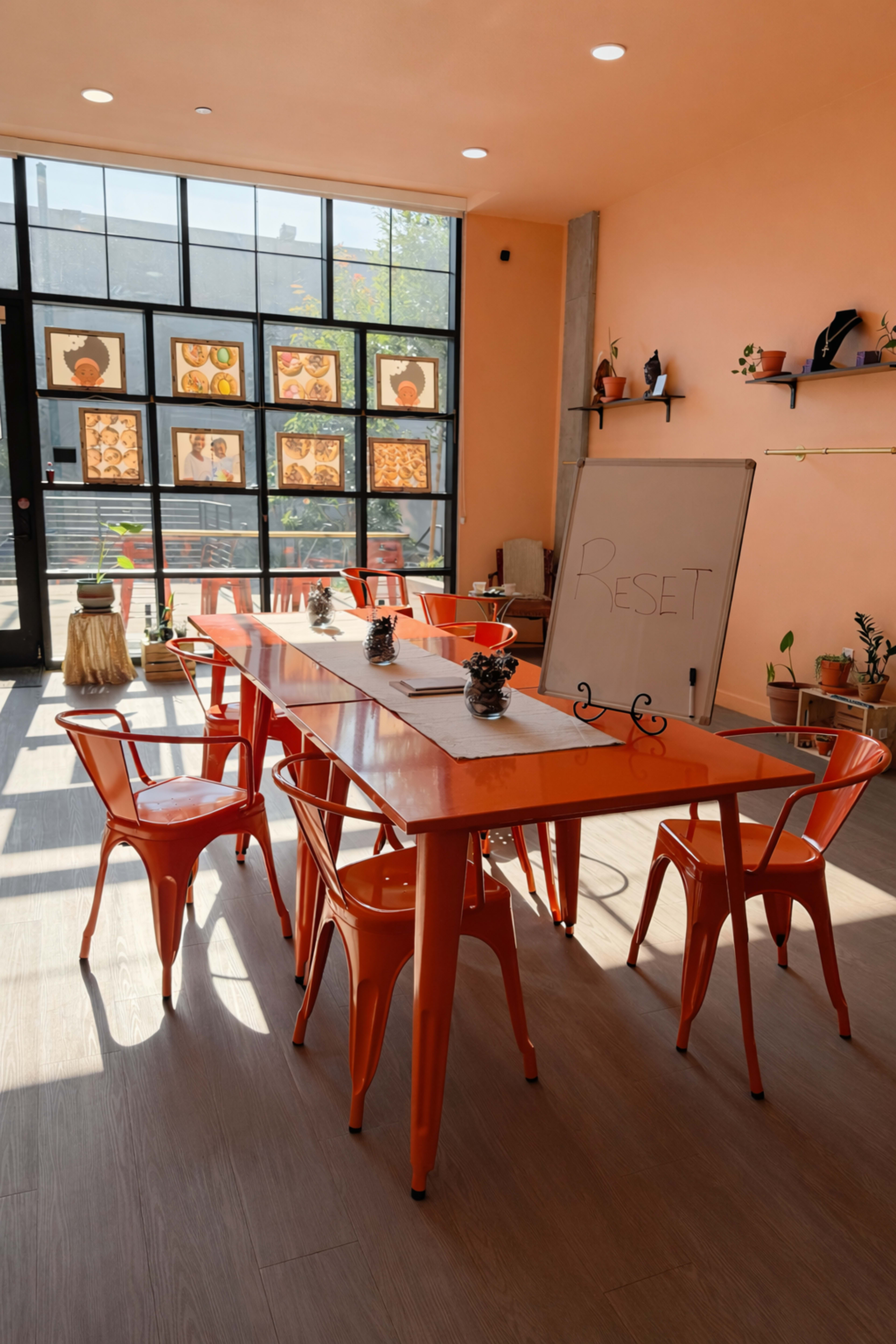 A spacious room features an orange table surrounded by matching chairs, with large windows displaying art and plants.