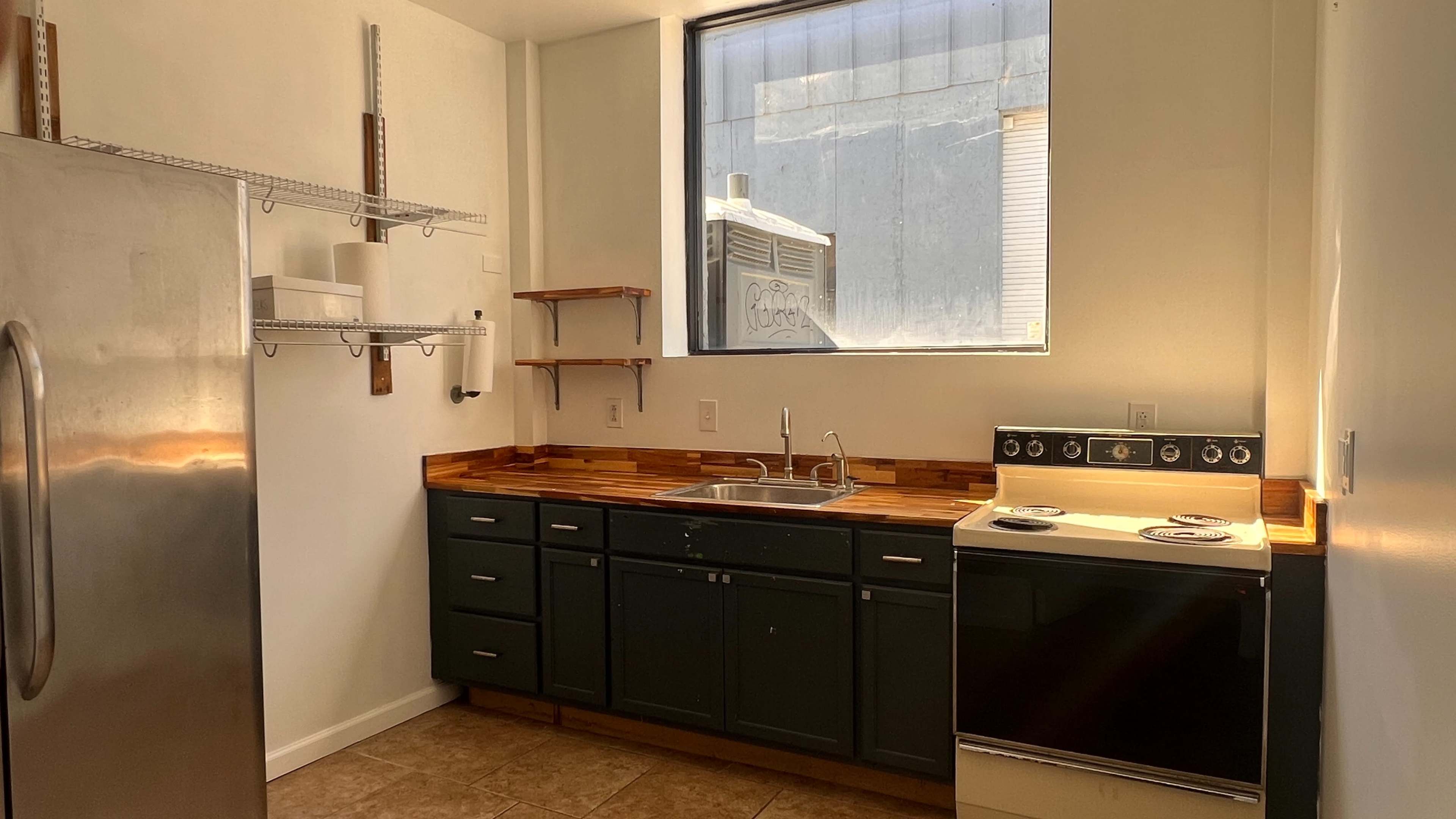 The image depicts a kitchen featuring gray cabinets, a stainless steel refrigerator, a stove, and a window that provides natural light.