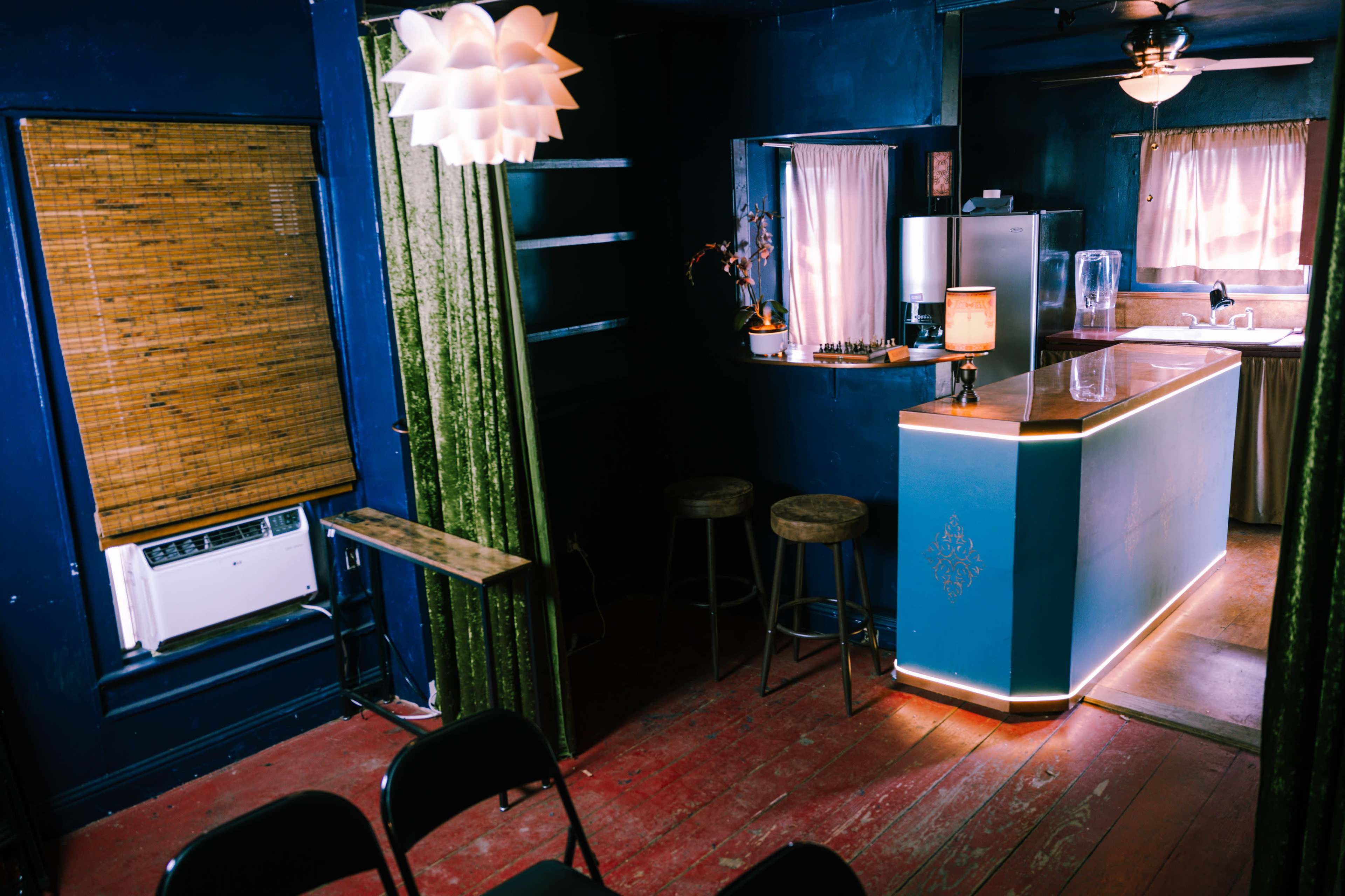 A cozy interior space featuring a bar with high stools, a window with bamboo blinds, and dark blue walls illuminated by a decorative light fixture.