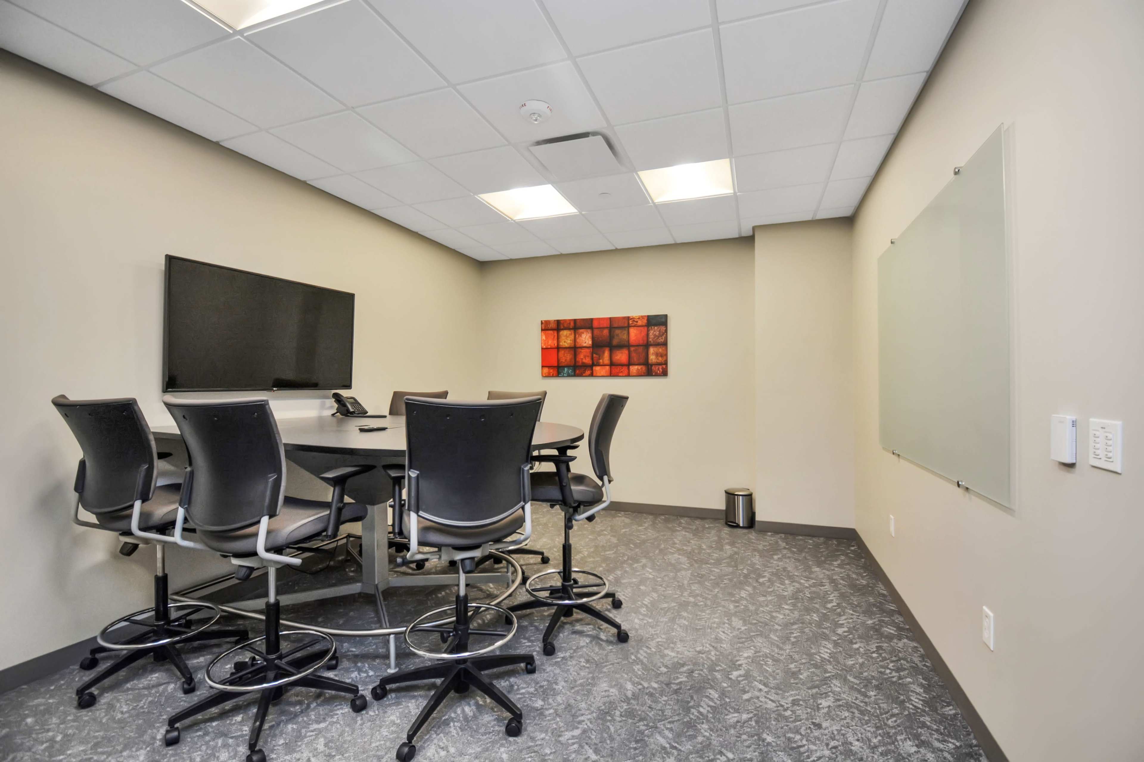 The image shows a modern conference room featuring a table surrounded by high-backed chairs, a wall-mounted television, and a whiteboard.