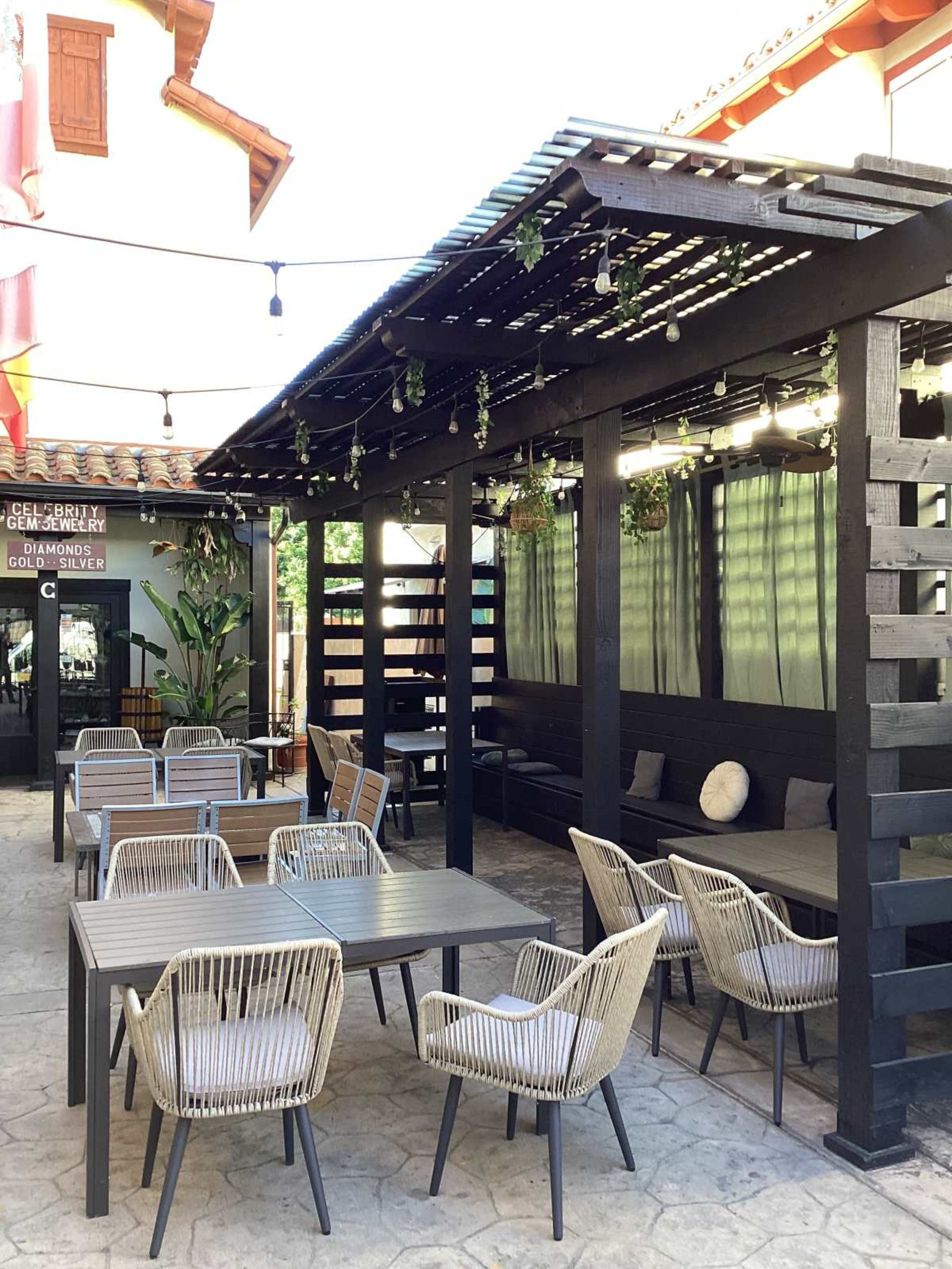 The image shows an outdoor dining area with several gray tables and woven chairs beneath a wooden pergola, adorned with string lights and greenery.