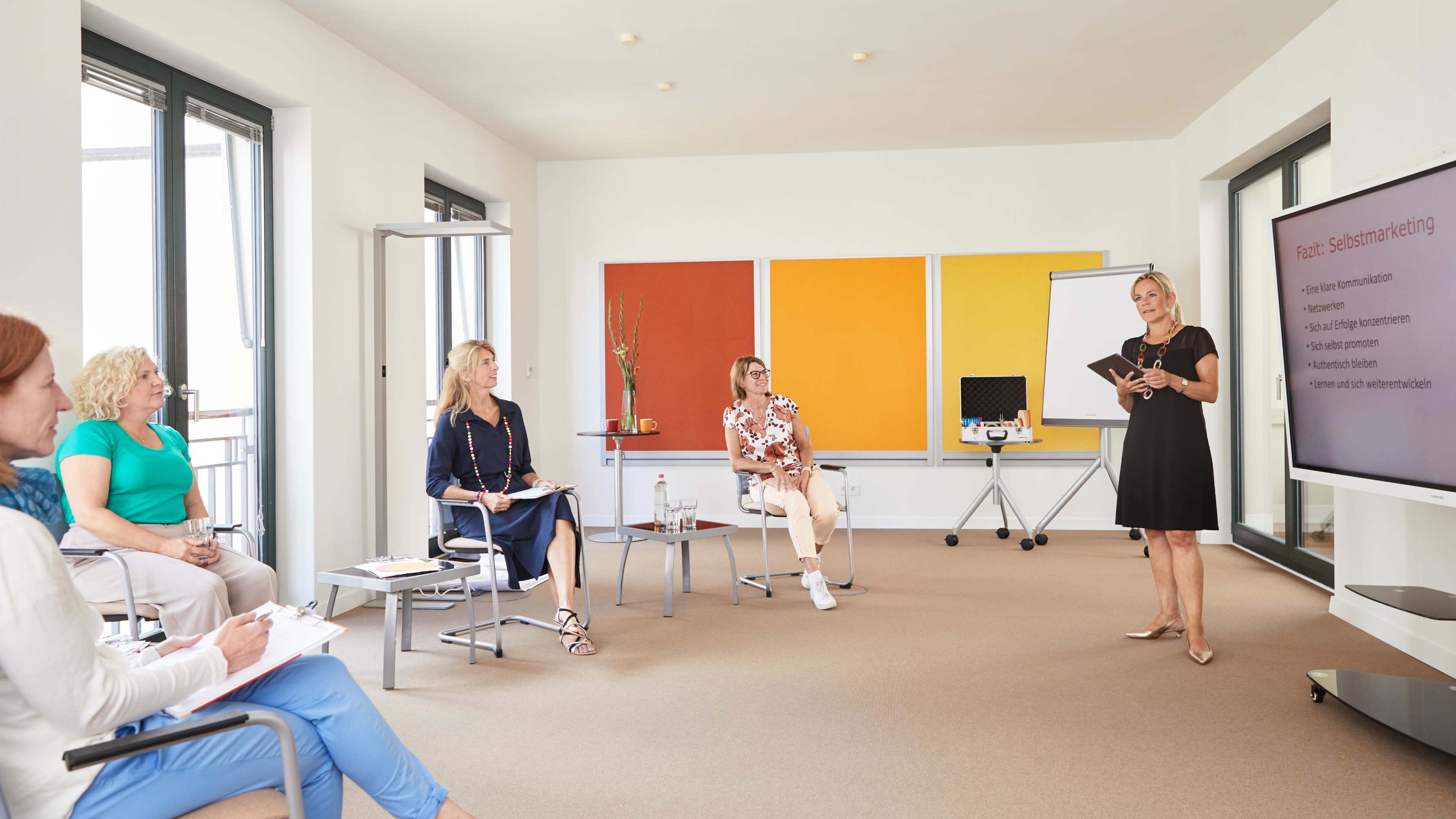 A woman stands at the front of a modern conference room presenting to a group of seated participants who are taking notes.