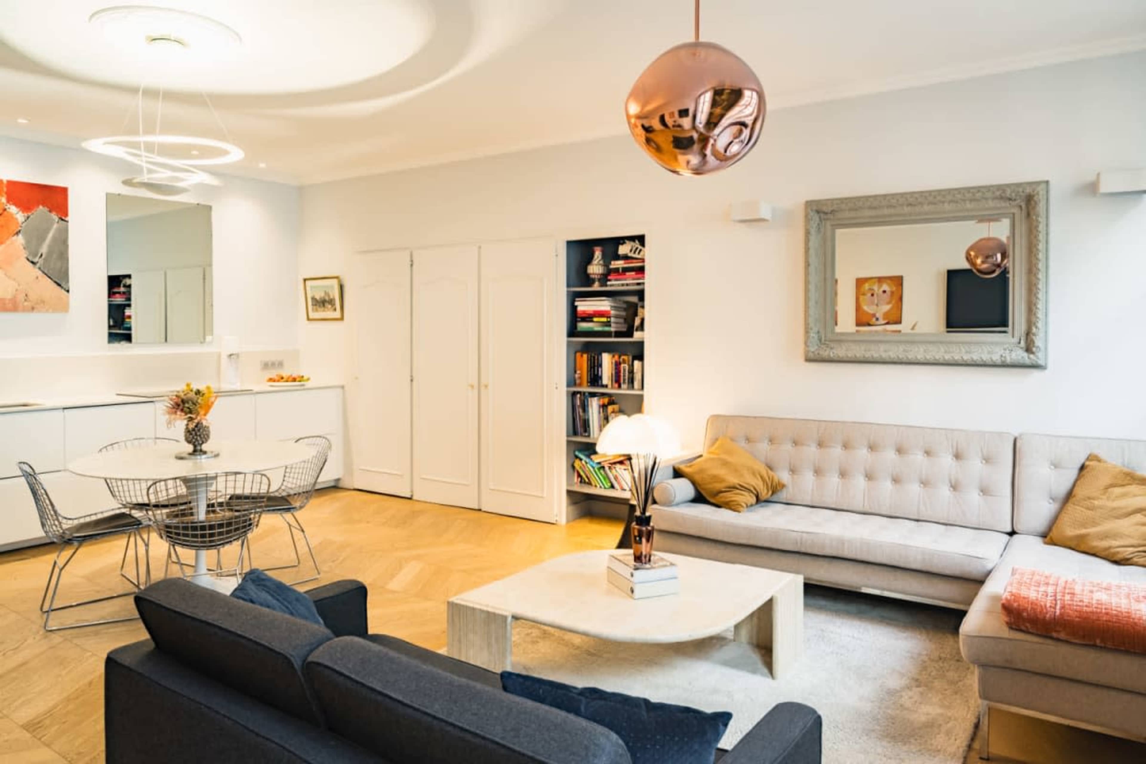 A modern living room features a light-colored sofa, a dark sofa, a round dining table with wire chairs, and a coffee table, all complemented by a mirrored wall and decorative lighting.
