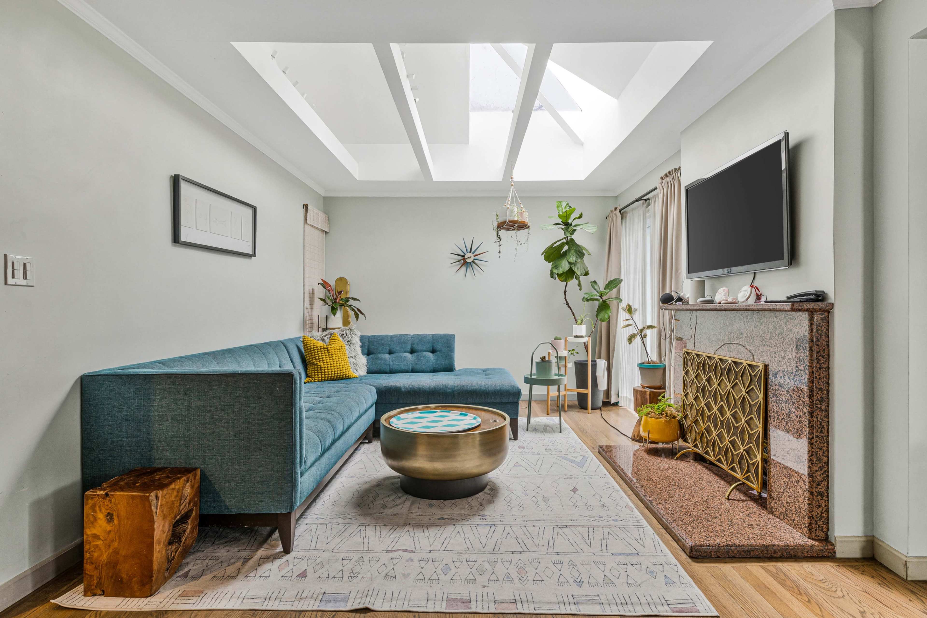 The living room features a blue sectional sofa, a gold coffee table, a television mounted on the wall, and a large skylight that illuminates the space.