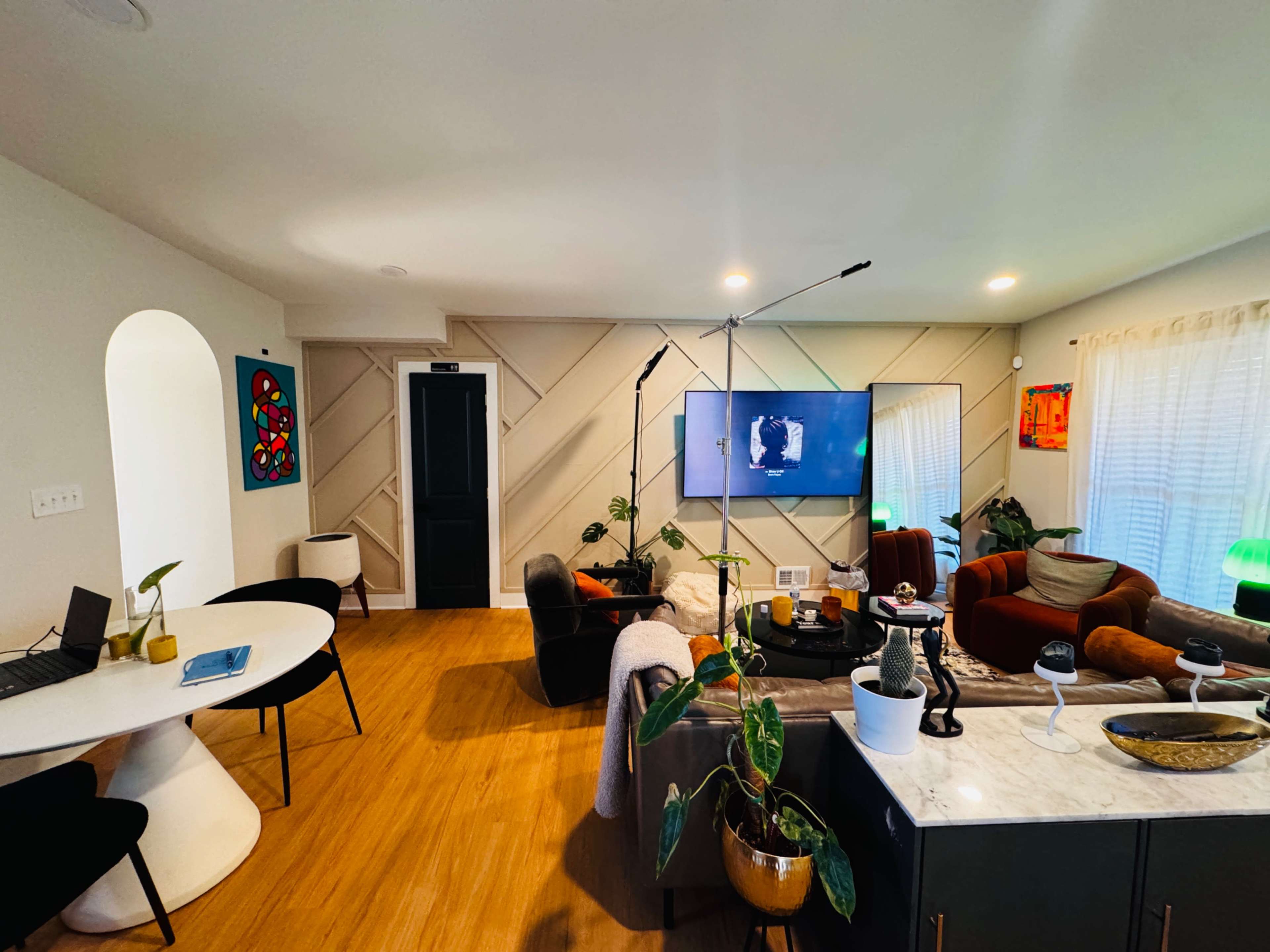 The image shows a modern living room with a central television, seating areas featuring a mix of colors, a dining table, and various plants.