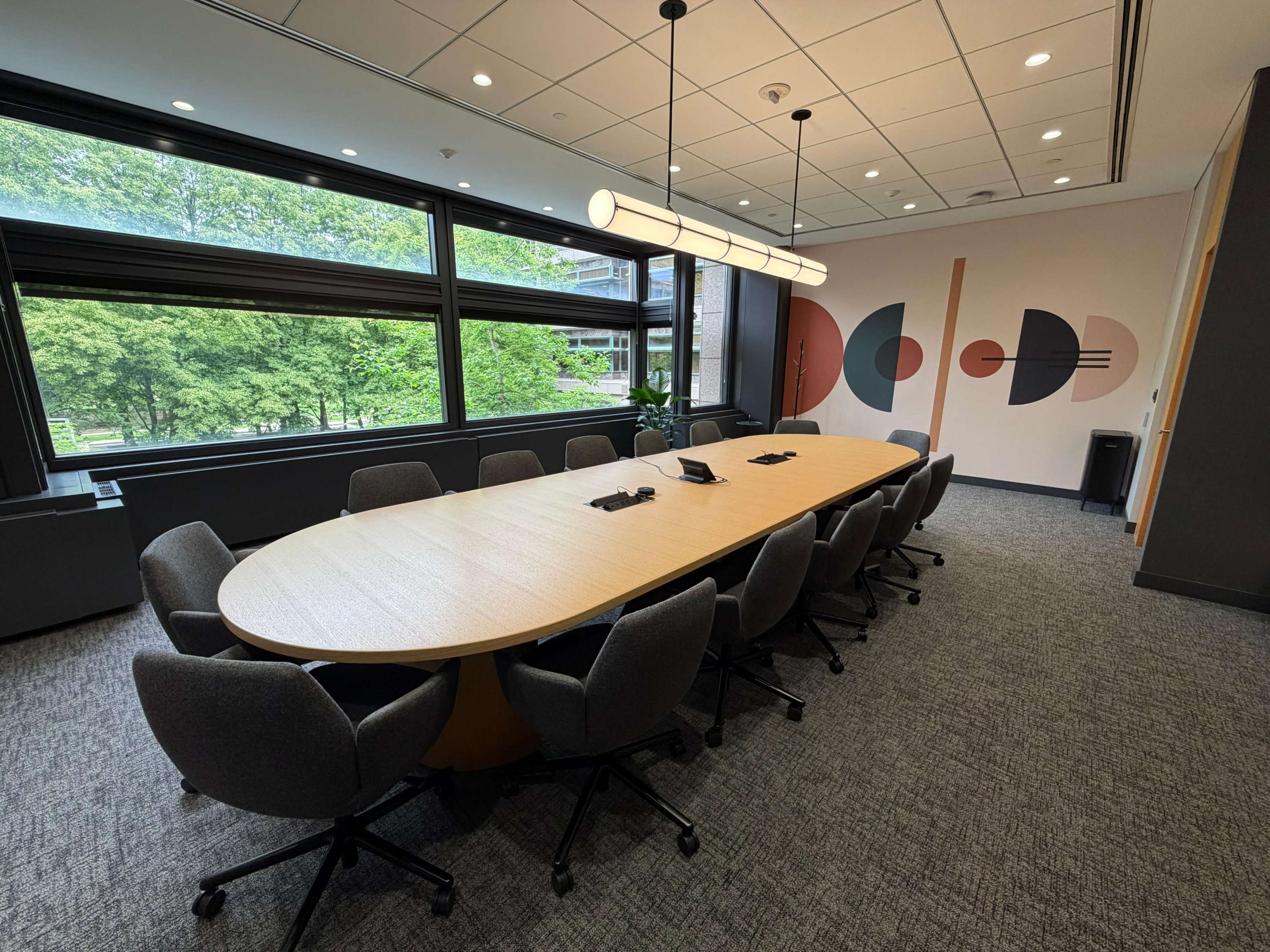 A modern conference room features a long oval table surrounded by ergonomic chairs and large windows overlooking greenery.