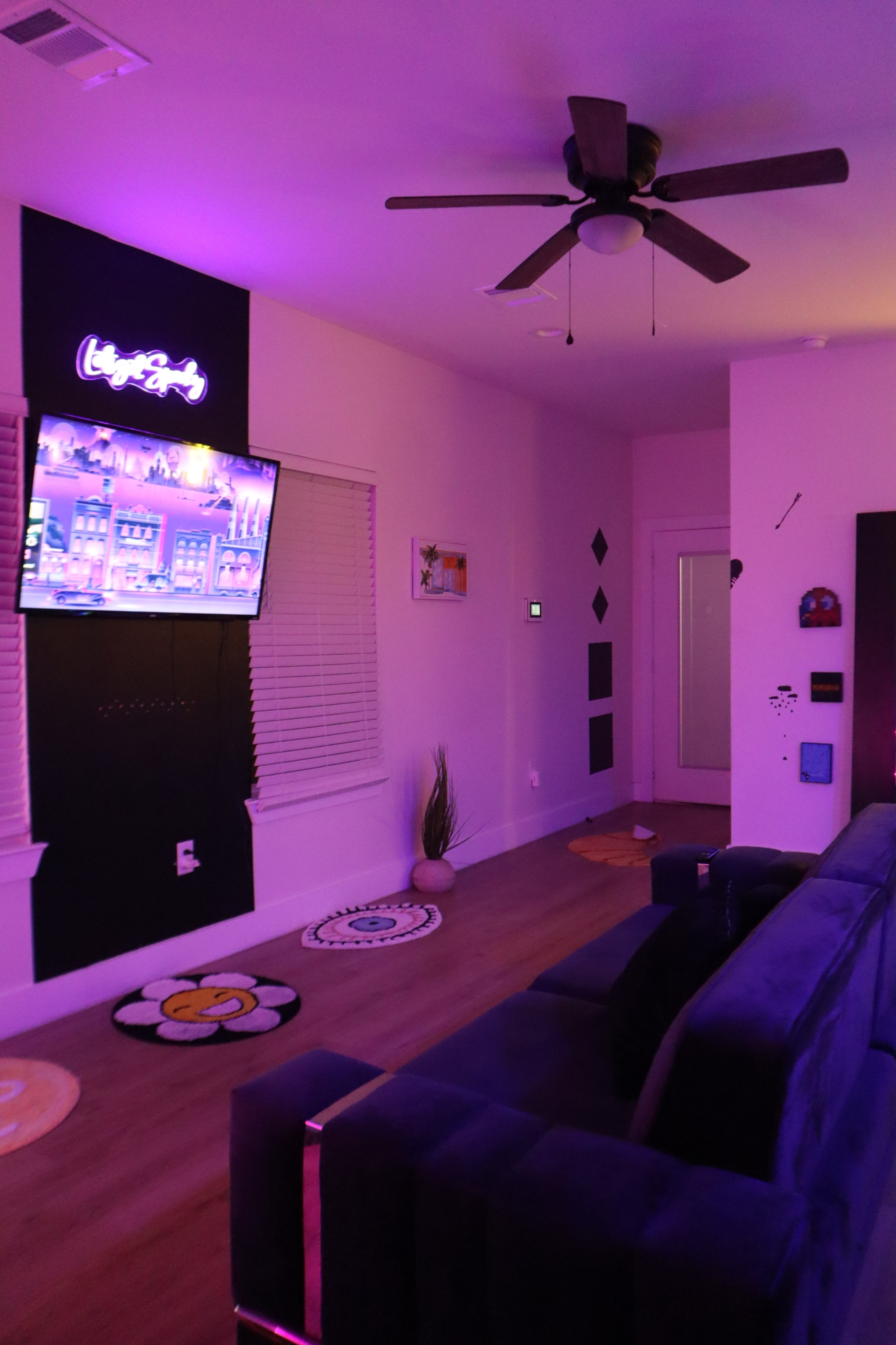 The room features a dark couch facing a wall-mounted television with a colorful display, illuminated by soft purple lighting.