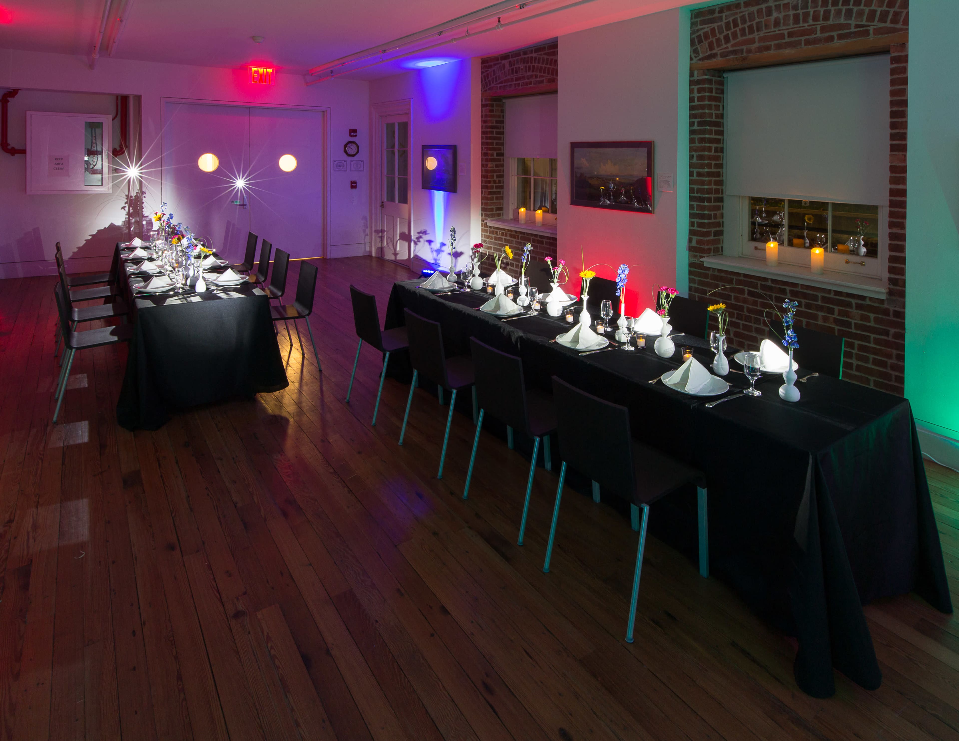 A long dining table is elegantly set with black linens, white dishes, and colorful floral centerpieces in a well-lit room with exposed brick walls.