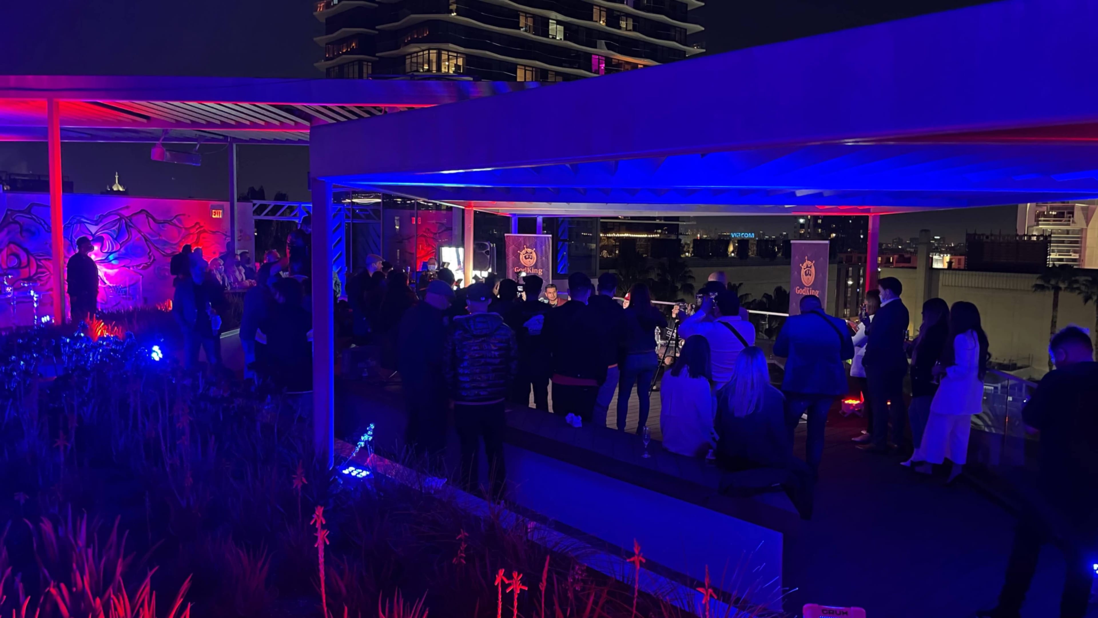 A crowd of people gathers in a brightly lit outdoor setting at night, with colorful lighting and modern architecture in the background.