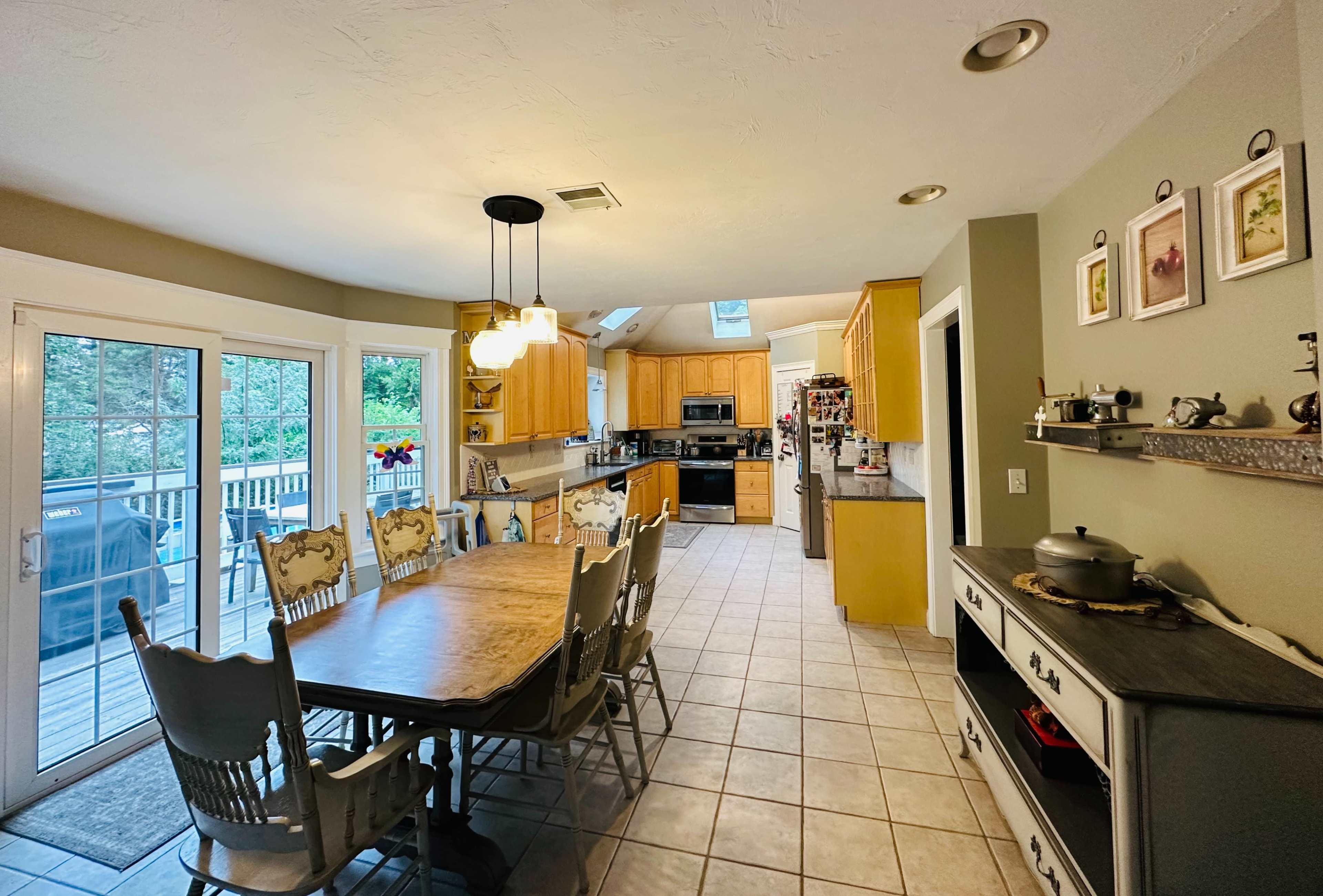 The image shows a spacious kitchen with wooden cabinets, a dining table surrounded by chairs, and large windows overlooking a deck.