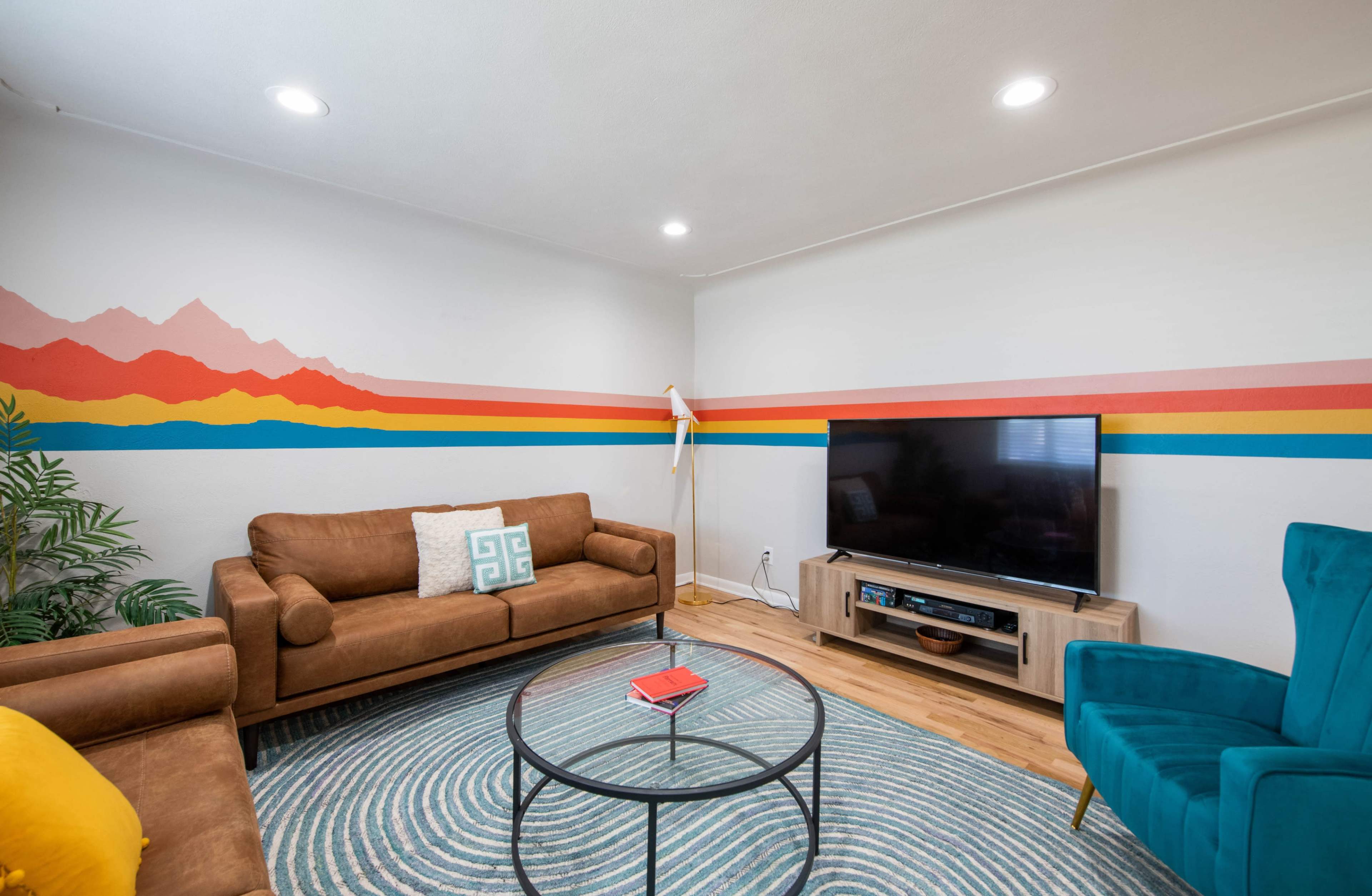 The living room features a brown leather sofa, a teal chair, a round coffee table, and a television mounted on a light wood console, all accented by colorful mountain and stripe murals on the wall.