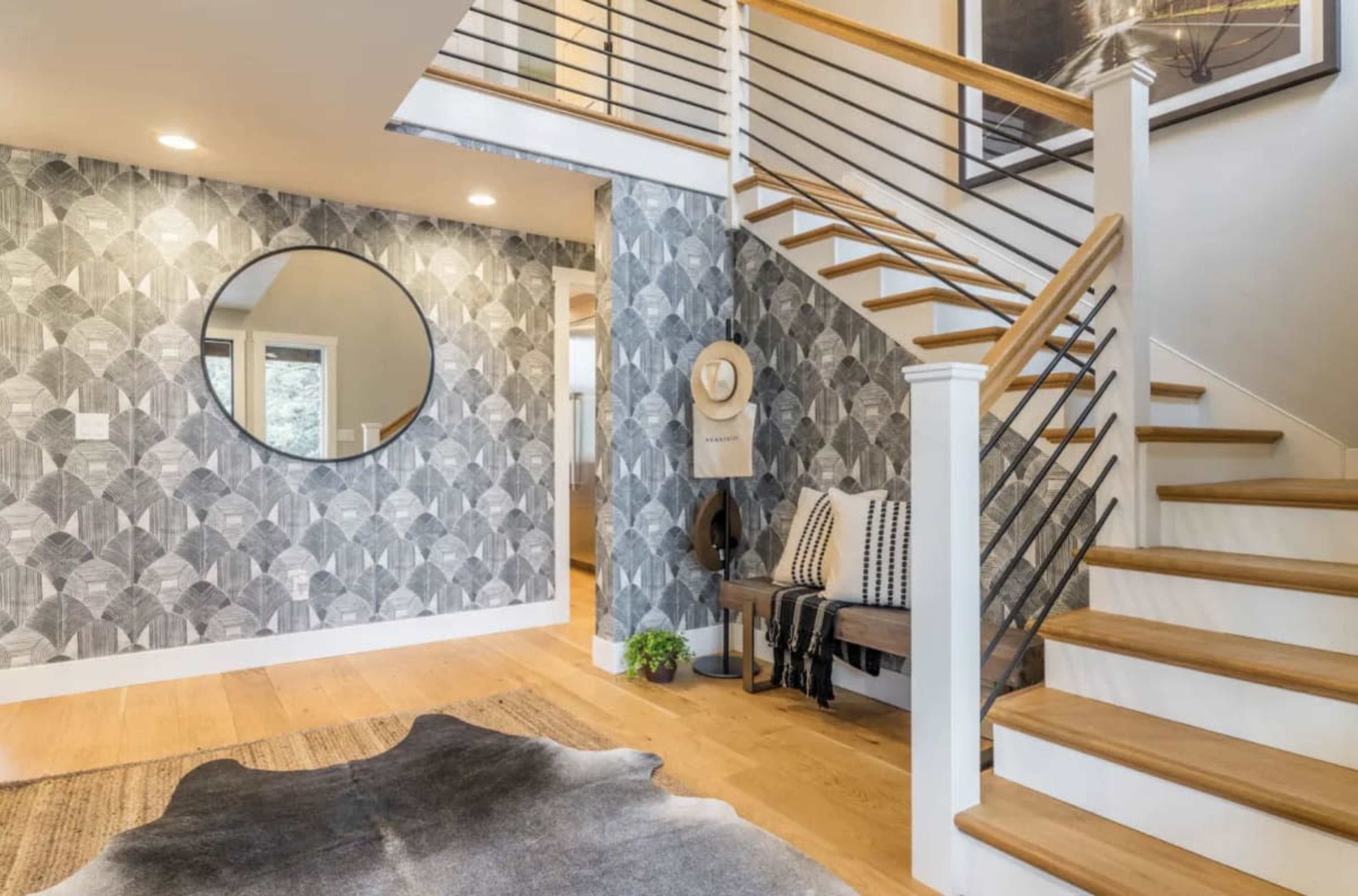 A modern entryway featuring a staircase with wooden steps, a geometric wallpaper design, a circular mirror, and a bench with decorative pillows.
