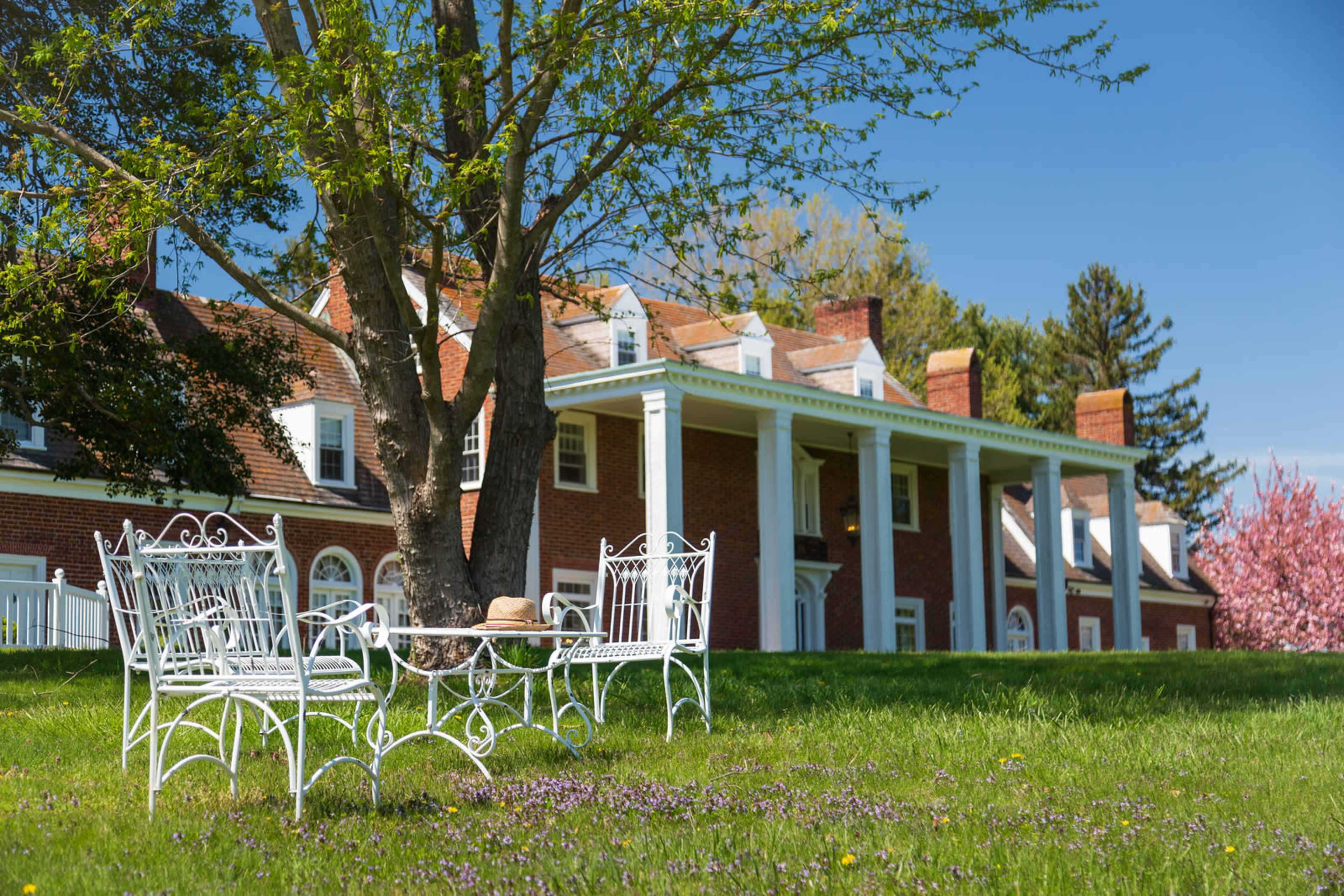 A white wrought iron table and chairs are placed under a tree in front of a large brick house with white columns.
