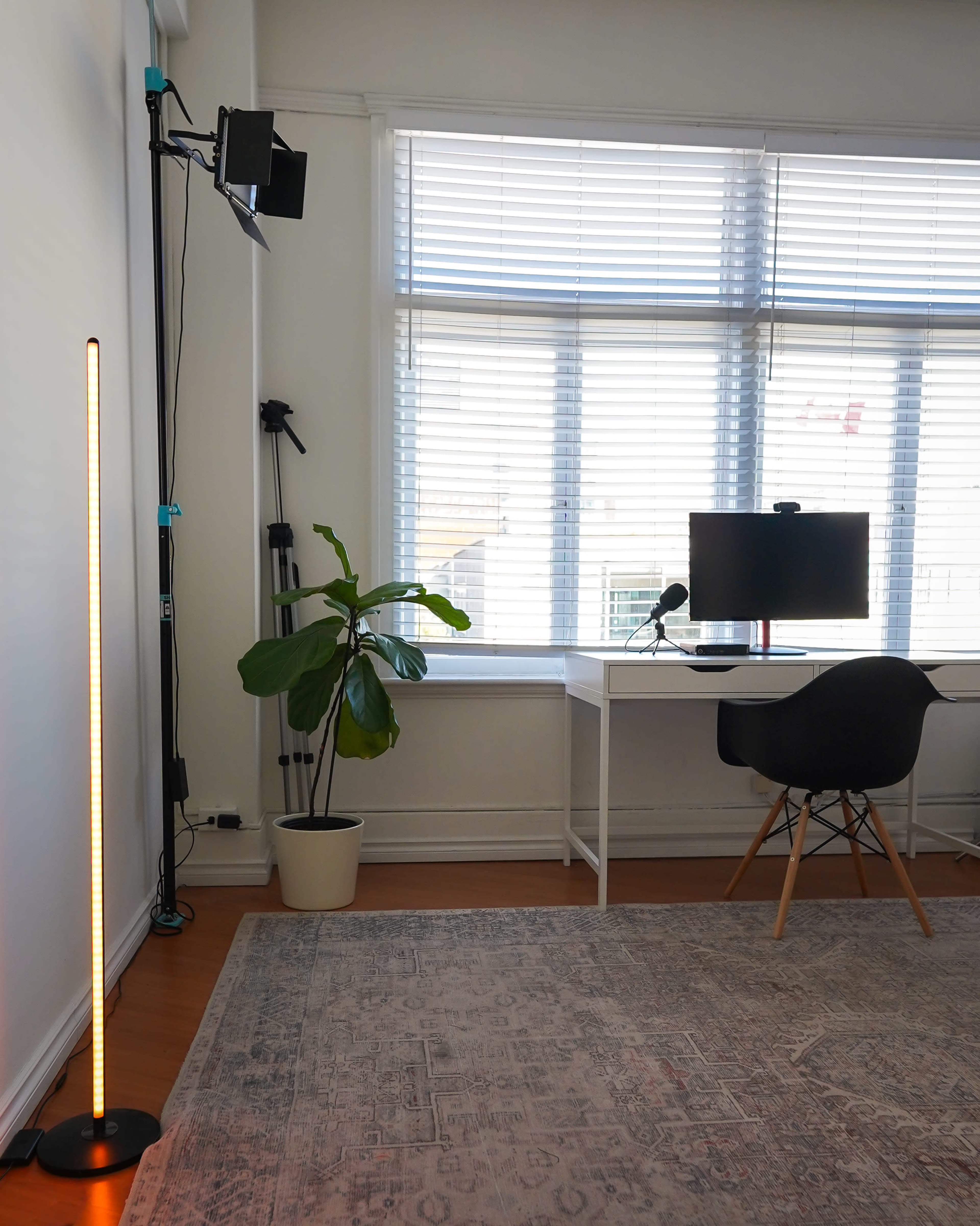 A bright, modern workspace featuring a desk with a monitor, a plant, and a tall floor lamp against a window with blinds.
