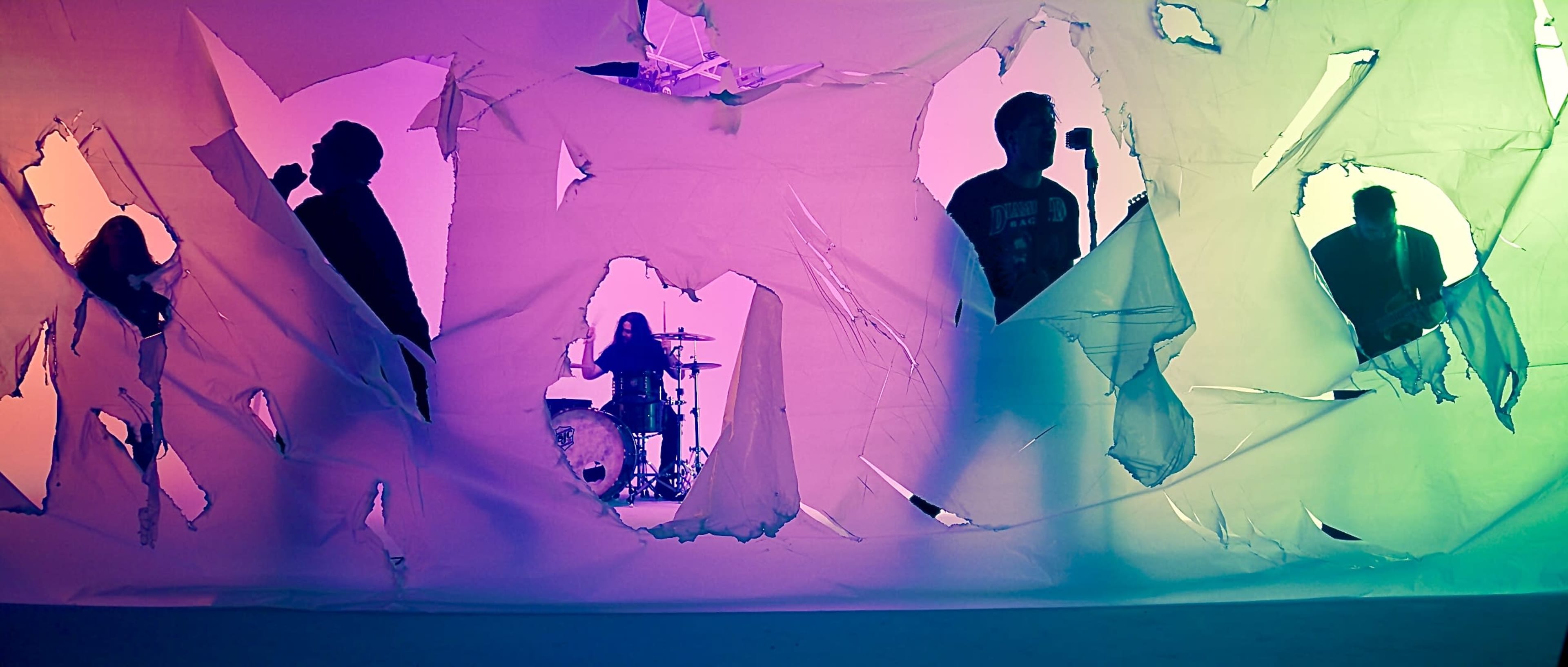 A band performs behind torn pieces of colorful backdrop, with a drummer visible in the center.