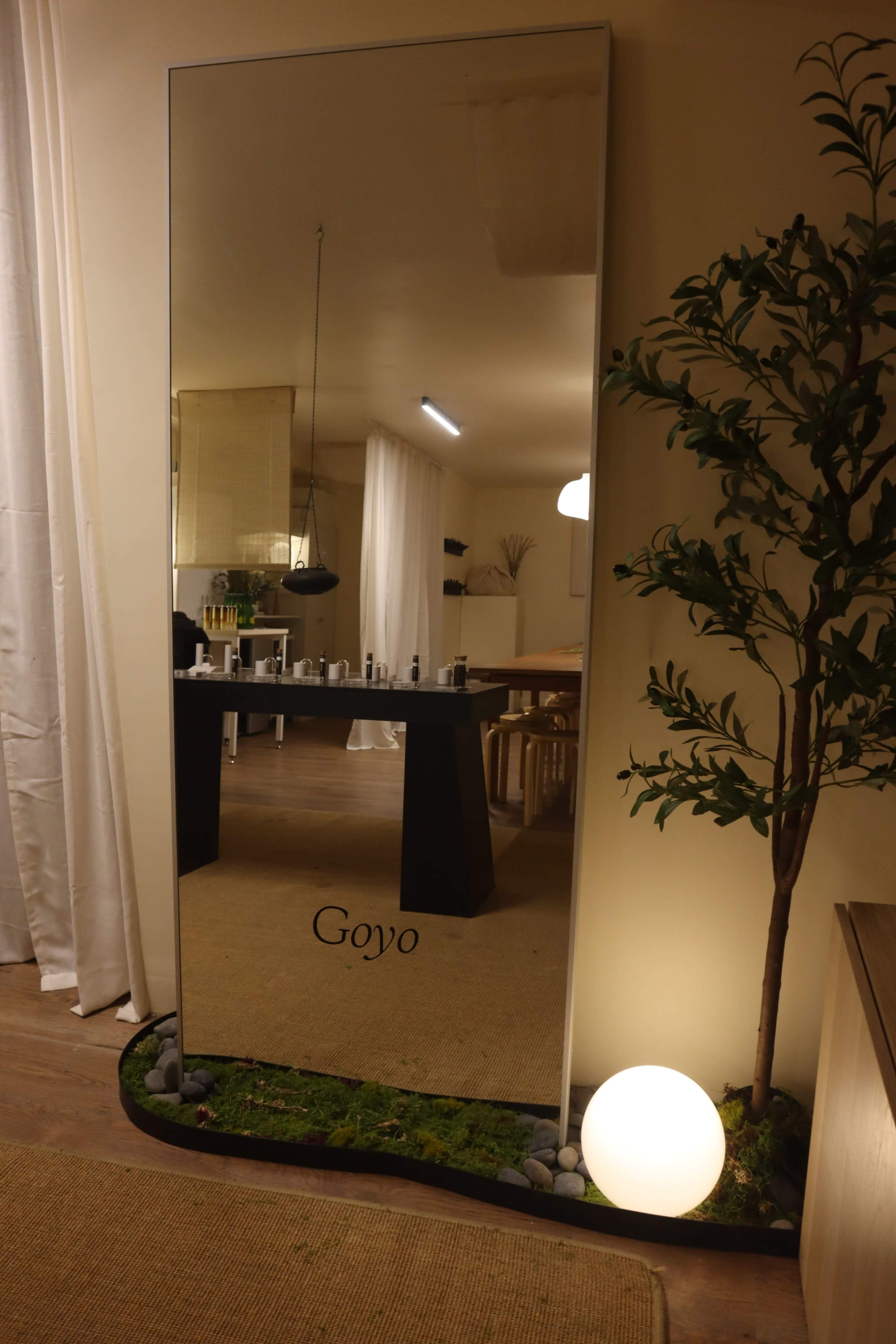 A large mirror with the name "Goyo" inscribed is positioned against a wall, reflecting a modern dining area with a black table and minimalistic decor.