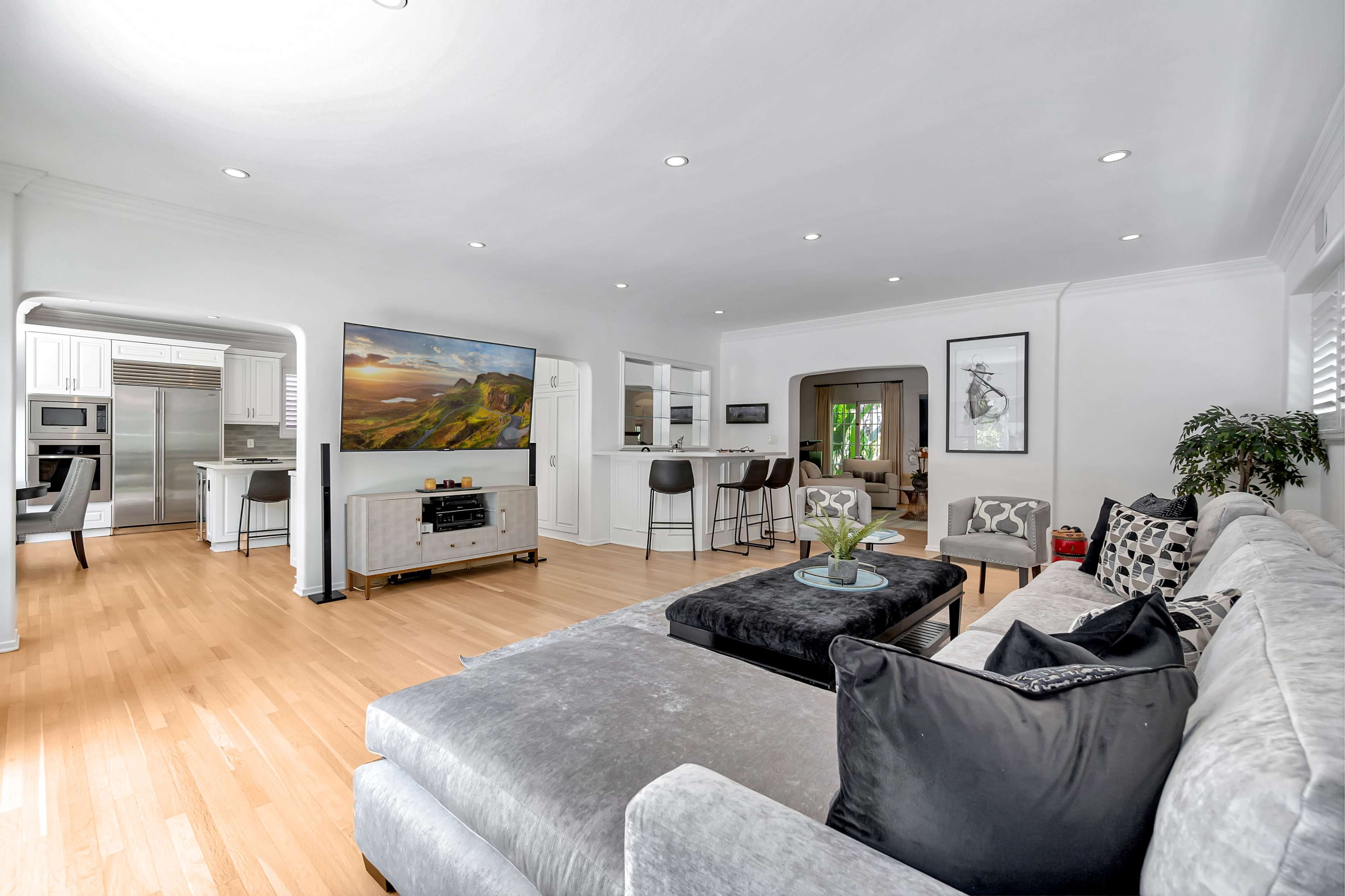 A modern living room features a sectional sofa, a coffee table, and a large television, with an open kitchen visible in the background.