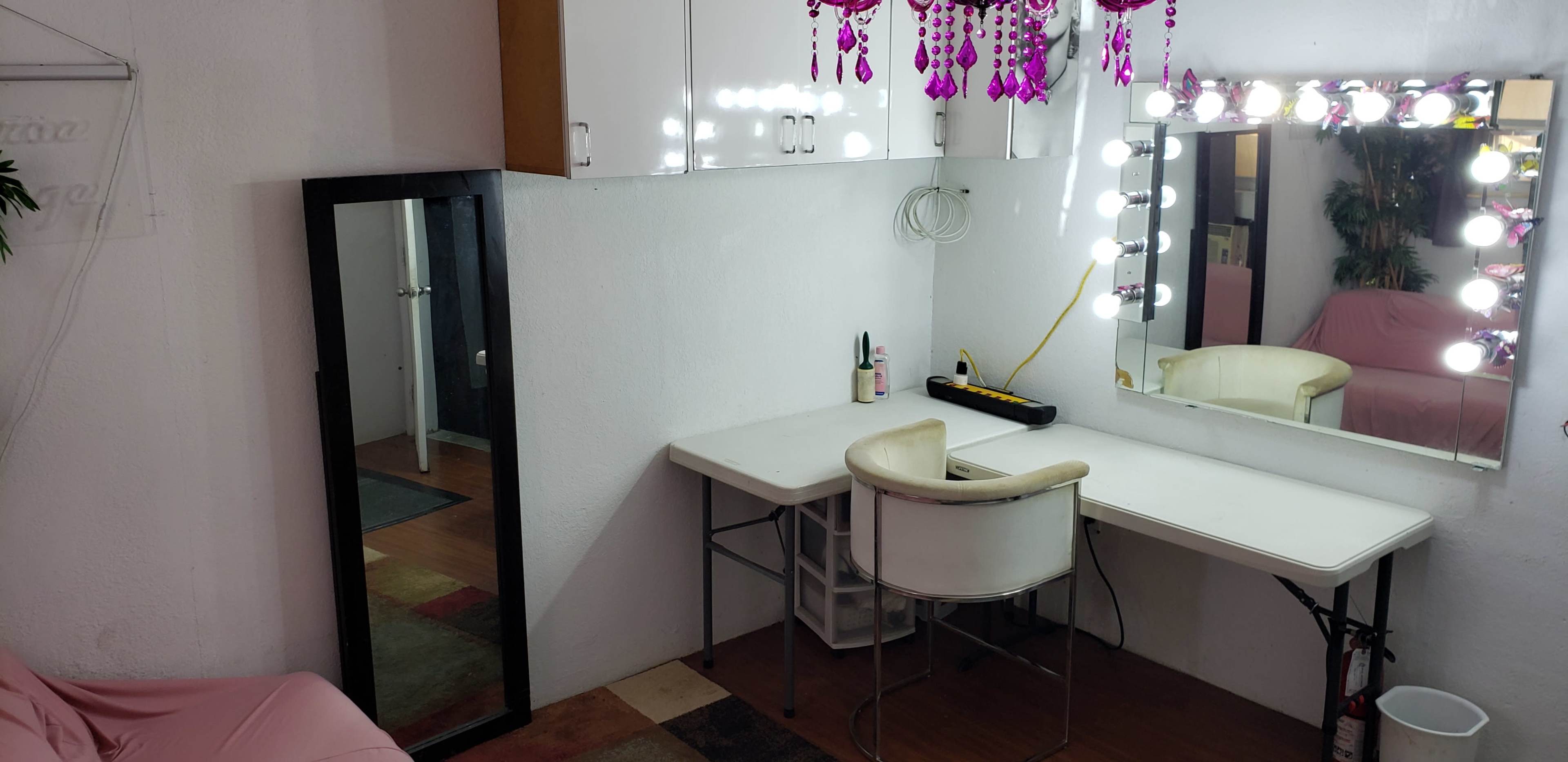A vanity area with a large mirror, bright lights, a white chair, and a decorative pink chandelier above.