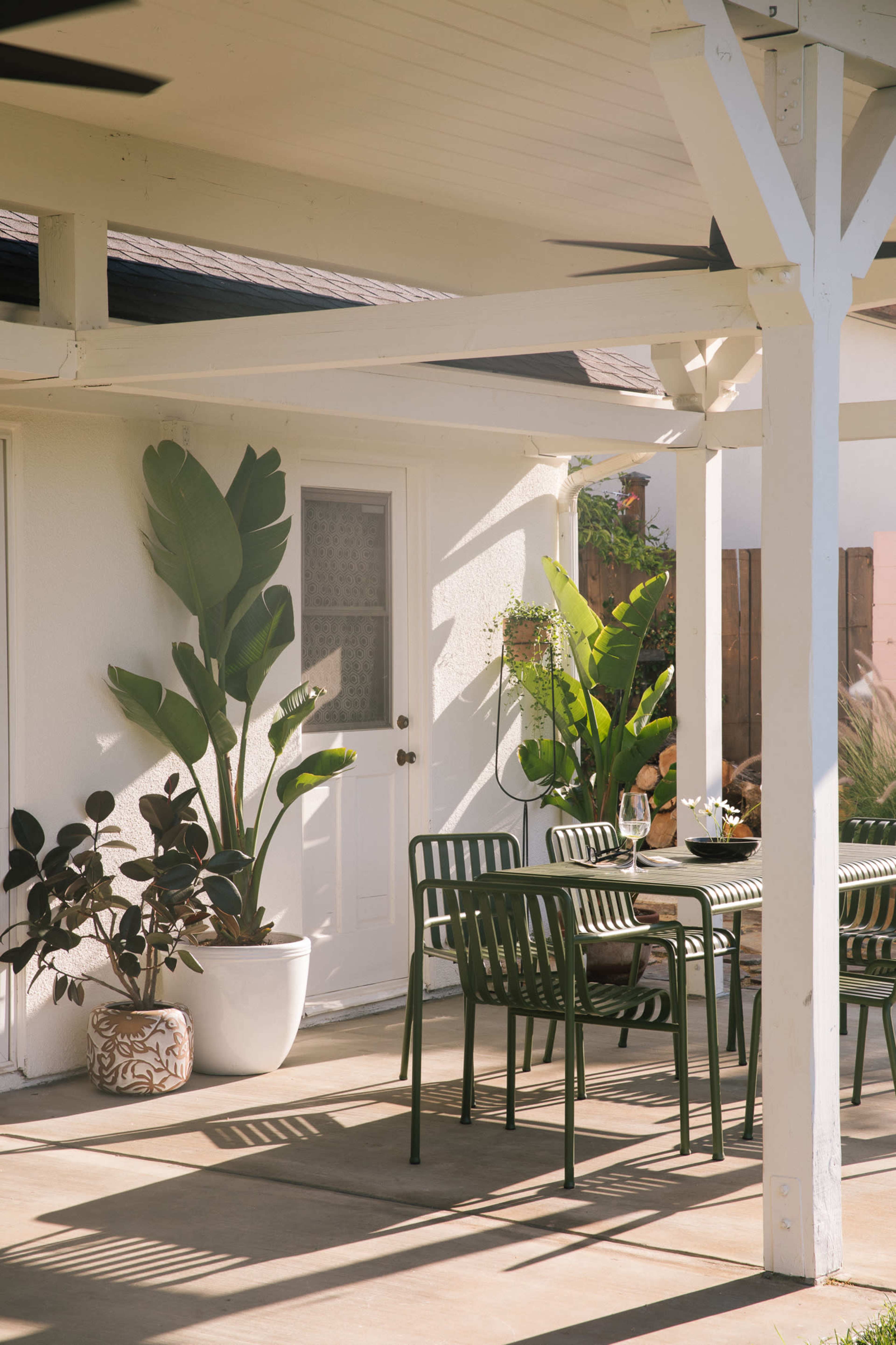 A patio area features a green dining set and potted plants under a white-beamed overhang.
