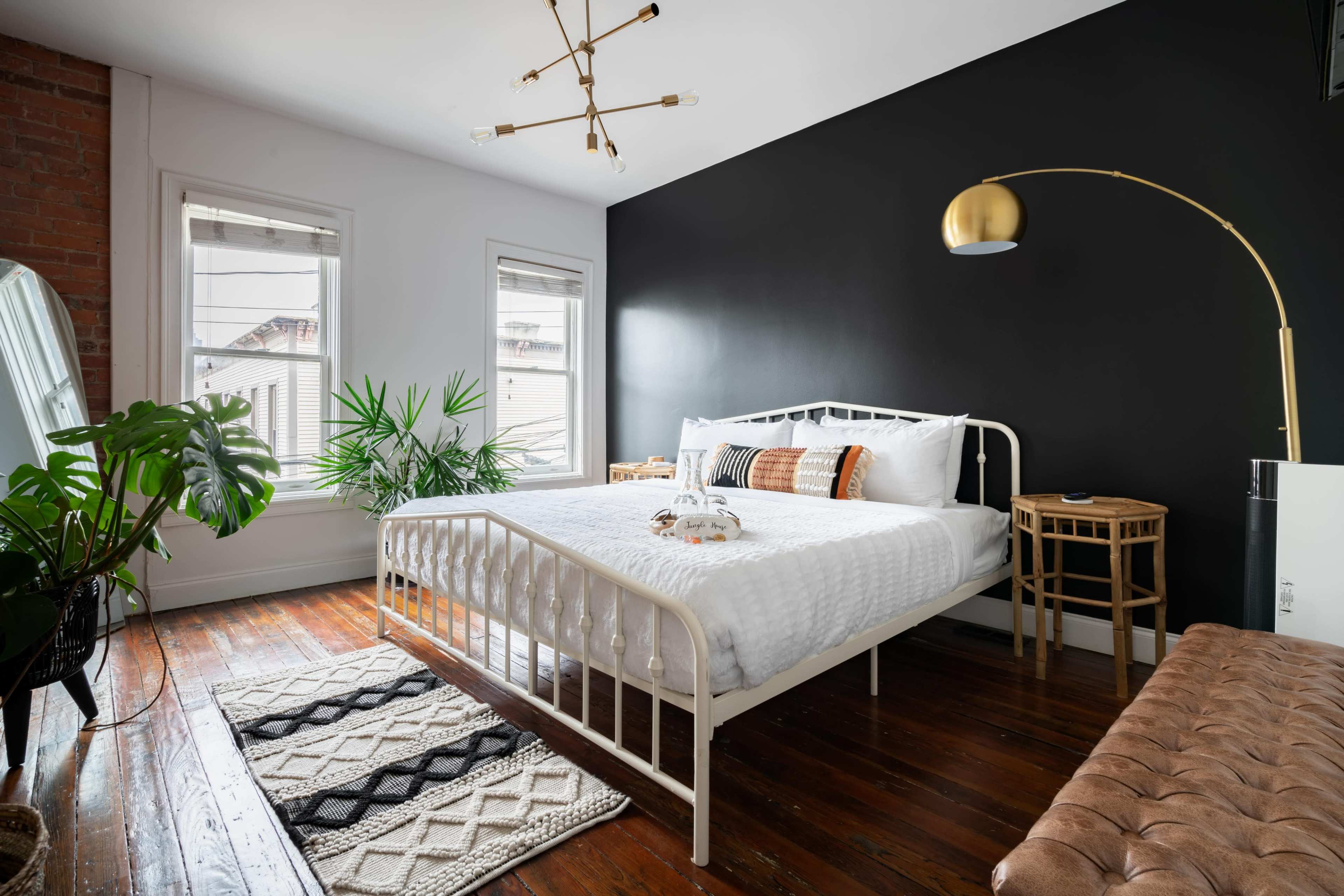 A bedroom features a white bed with decorative pillows, a wood floor, a large floor lamp, and a wall painted black.