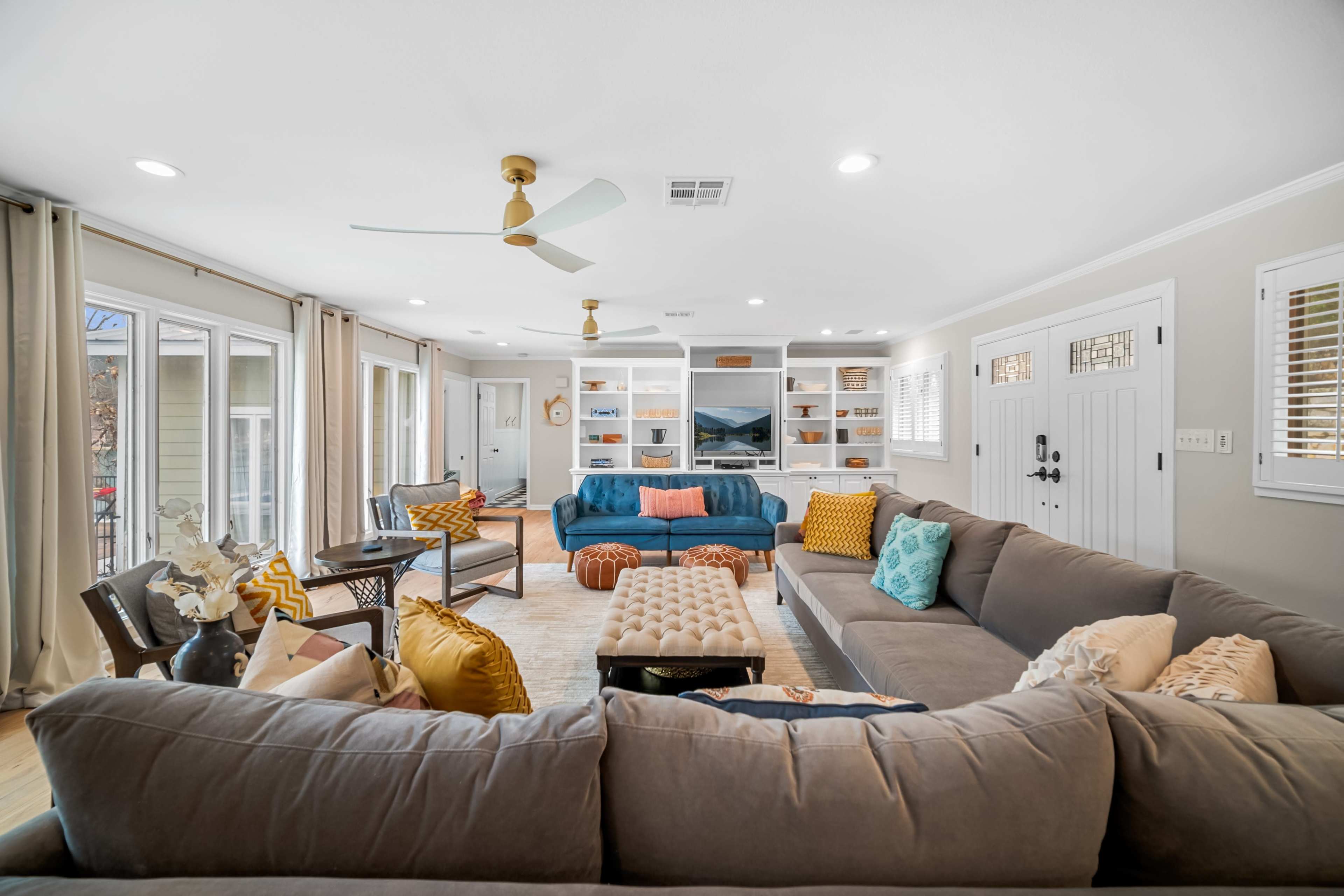 The living room features a sectional sofa, vibrant throw pillows, a coffee table, and built-in shelves, with large windows allowing natural light.