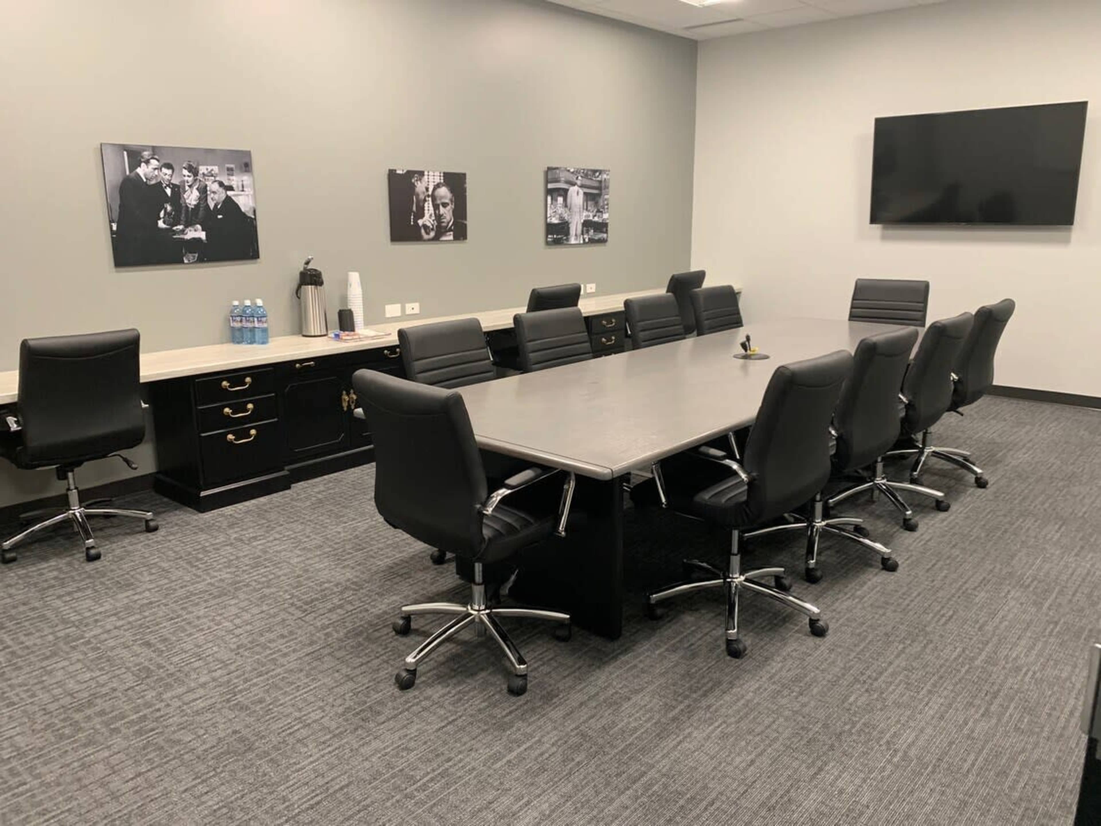 A modern conference room with a long table surrounded by black swivel chairs, a large screen on the wall, and water bottles on a counter in the background.