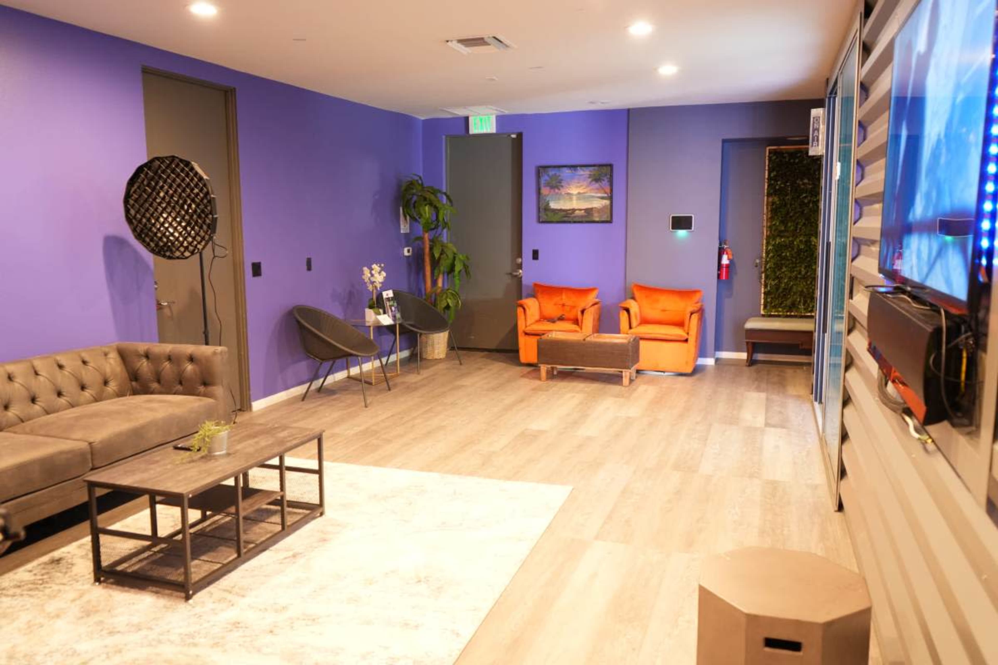 The image shows a modern lounge area featuring a brown tufted sofa, orange chairs, a coffee table, and purple walls with decorative plants.
