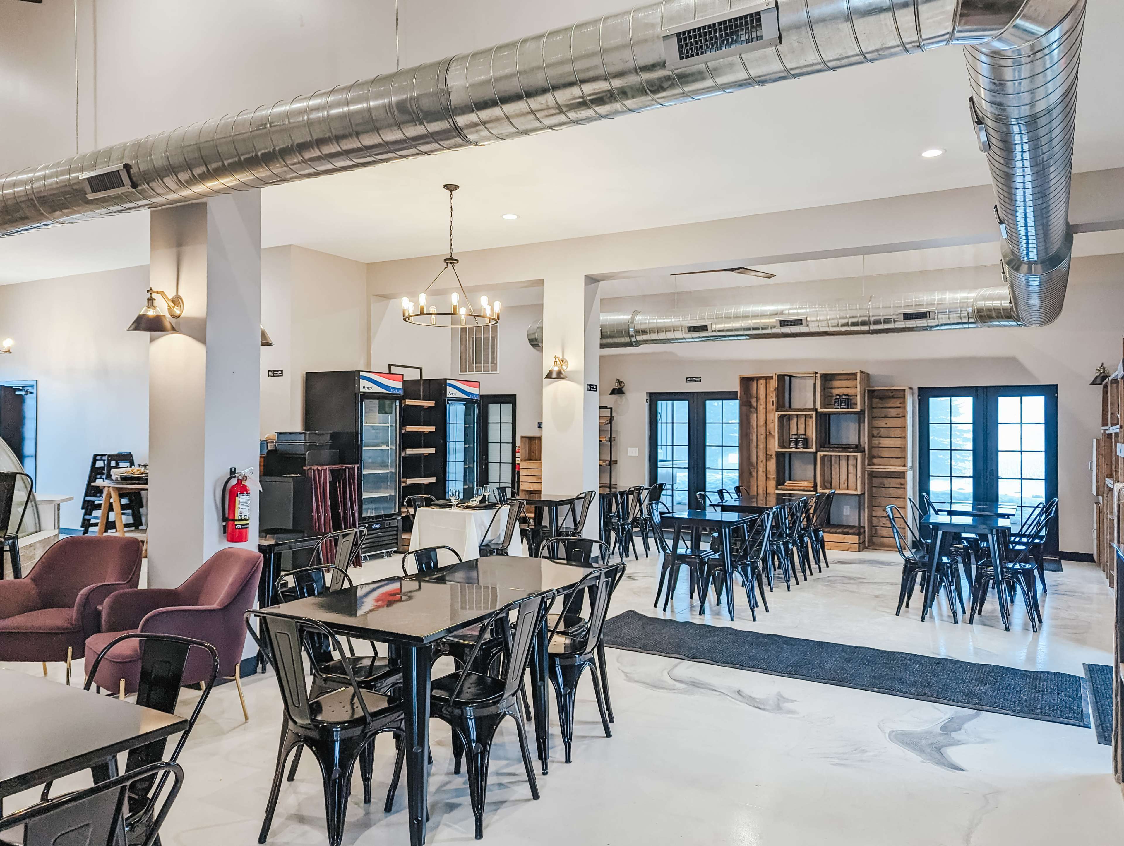 A spacious, modern café interior with black metal tables and chairs, purple armchairs, and large windows, featuring industrial-style ducts along the ceiling.