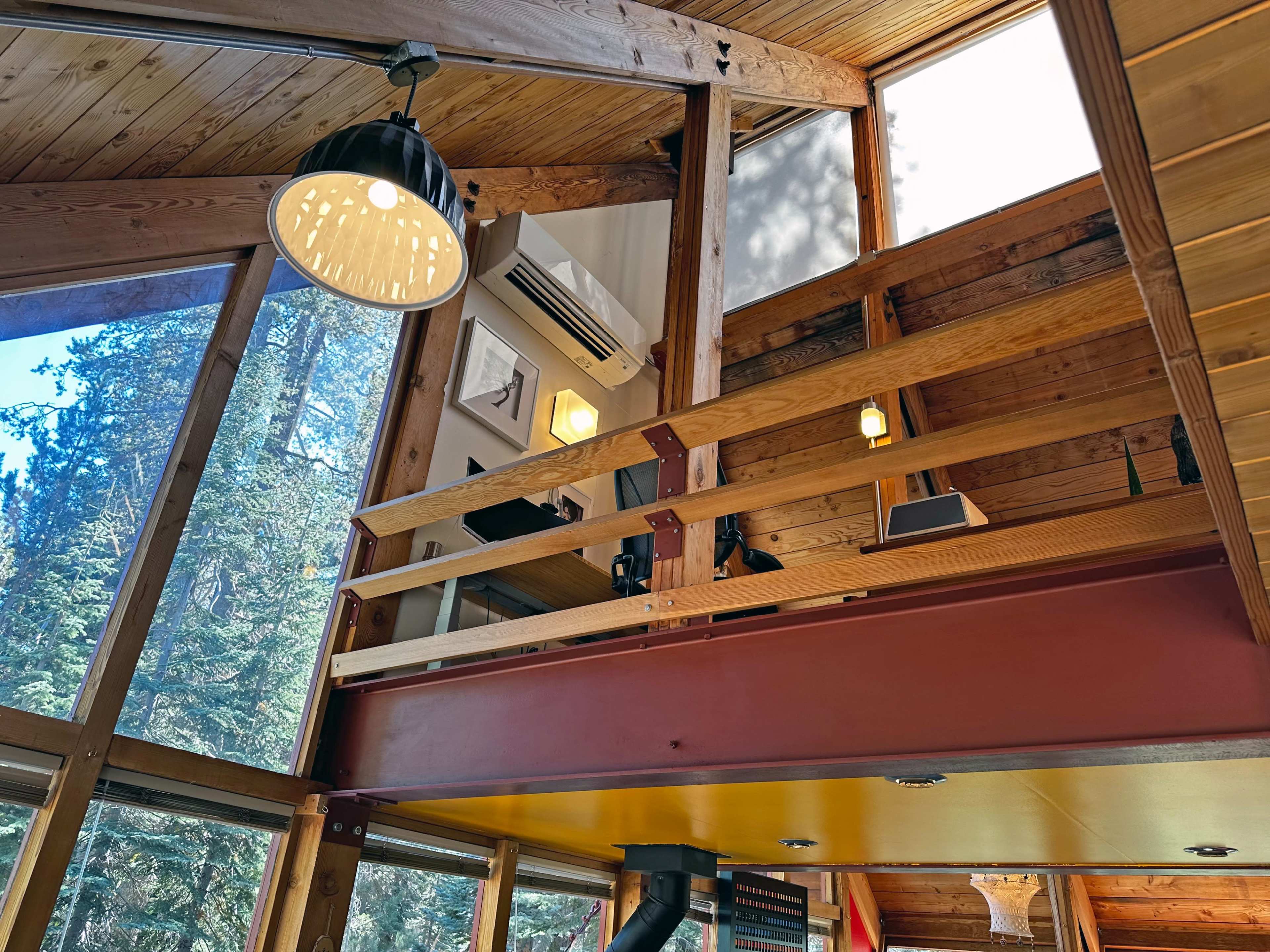 The image shows the interior of a wooden cabin with a loft area, large windows allowing natural light, and various decorative elements on the walls.