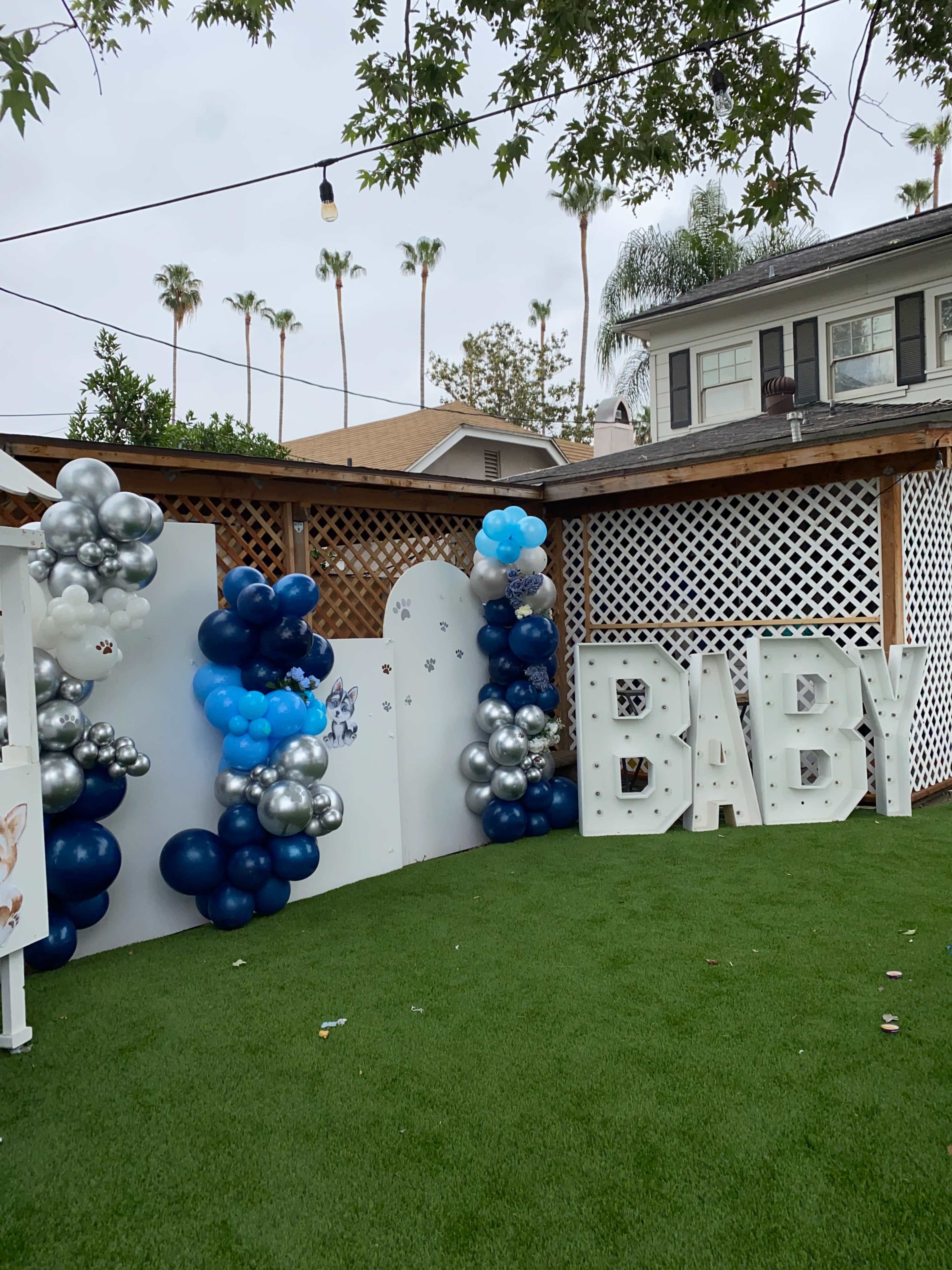 The scene features a decorated outdoor area with a green lawn, a backdrop of white balloons in blue and silver, and large white letters spelling "BABY."