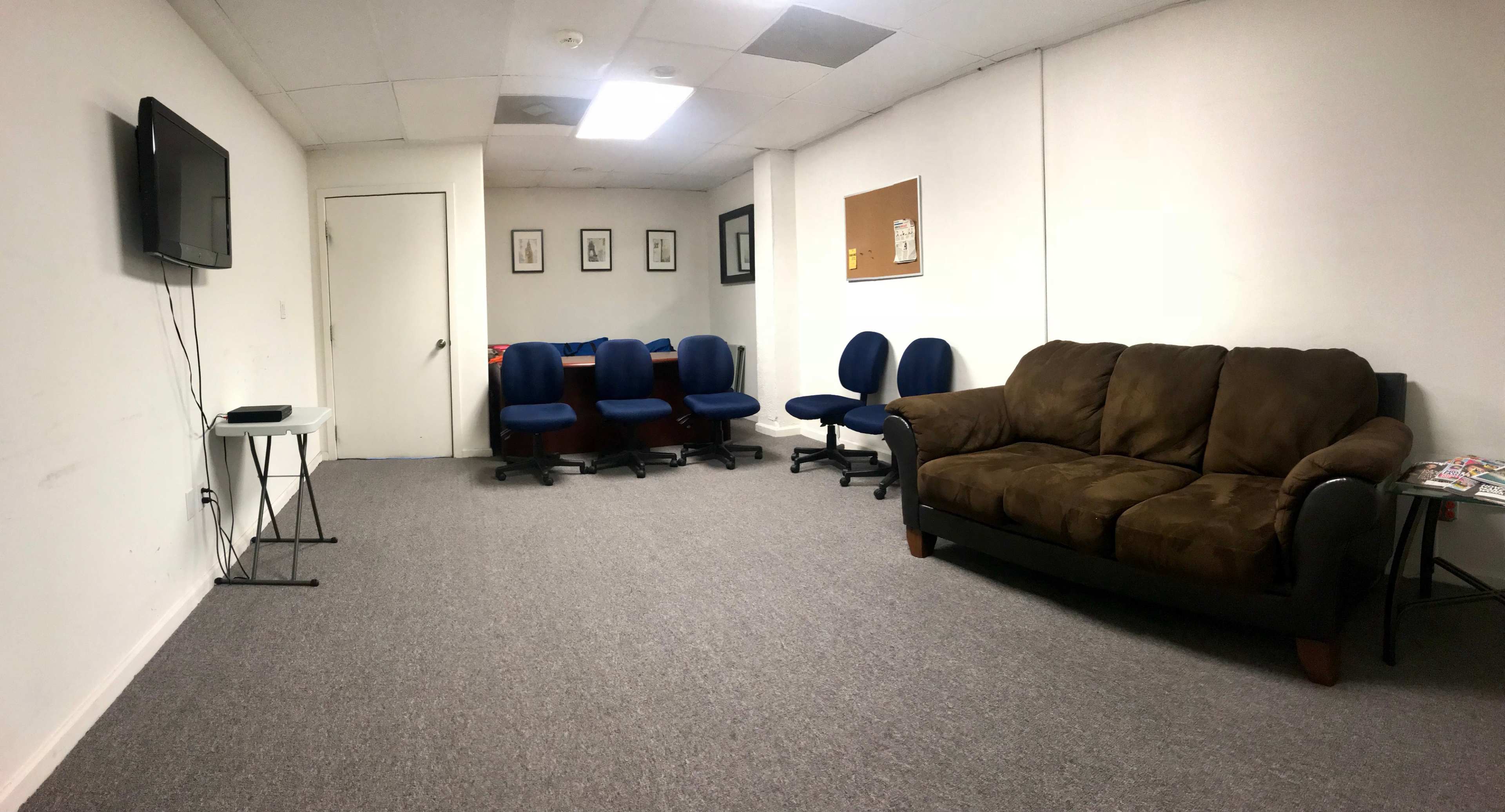 A sparsely furnished room with a brown couch, several blue chairs arranged around a table, a television on the wall, and a bulletin board.