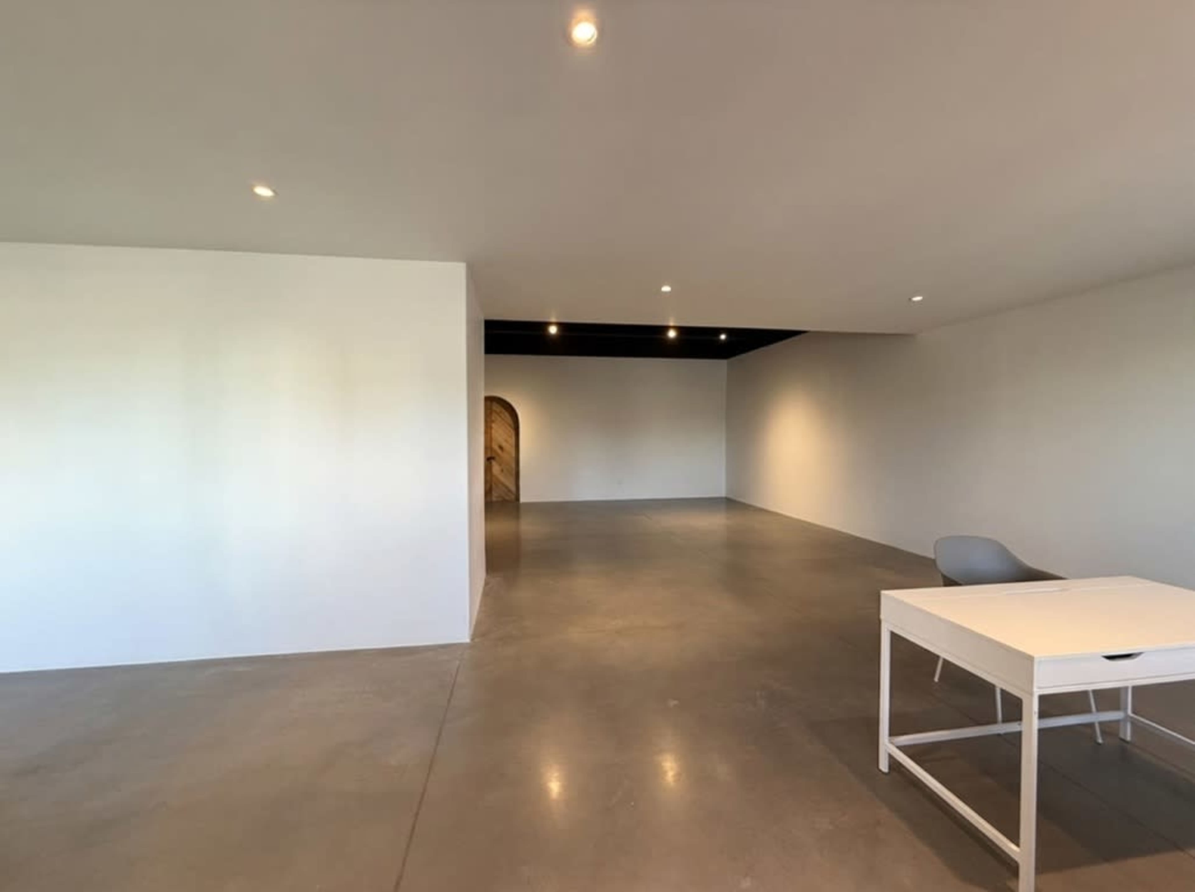 The image shows a spacious, empty room with a concrete floor, a white wall, and a desk with a chair in one corner.