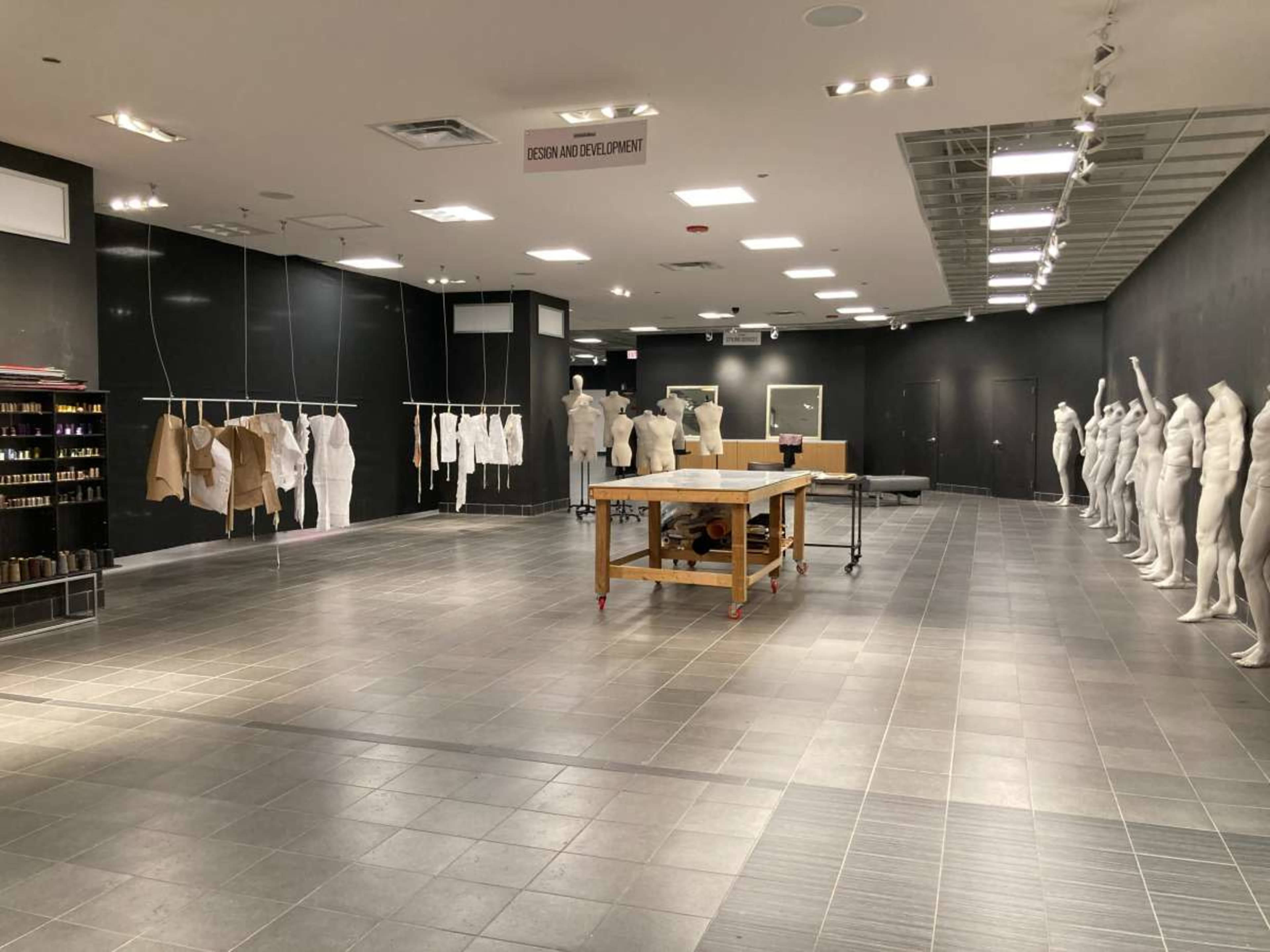 The image shows a spacious interior room with several clothing mannequins and a central work table, surrounded by black walls and tiled flooring.