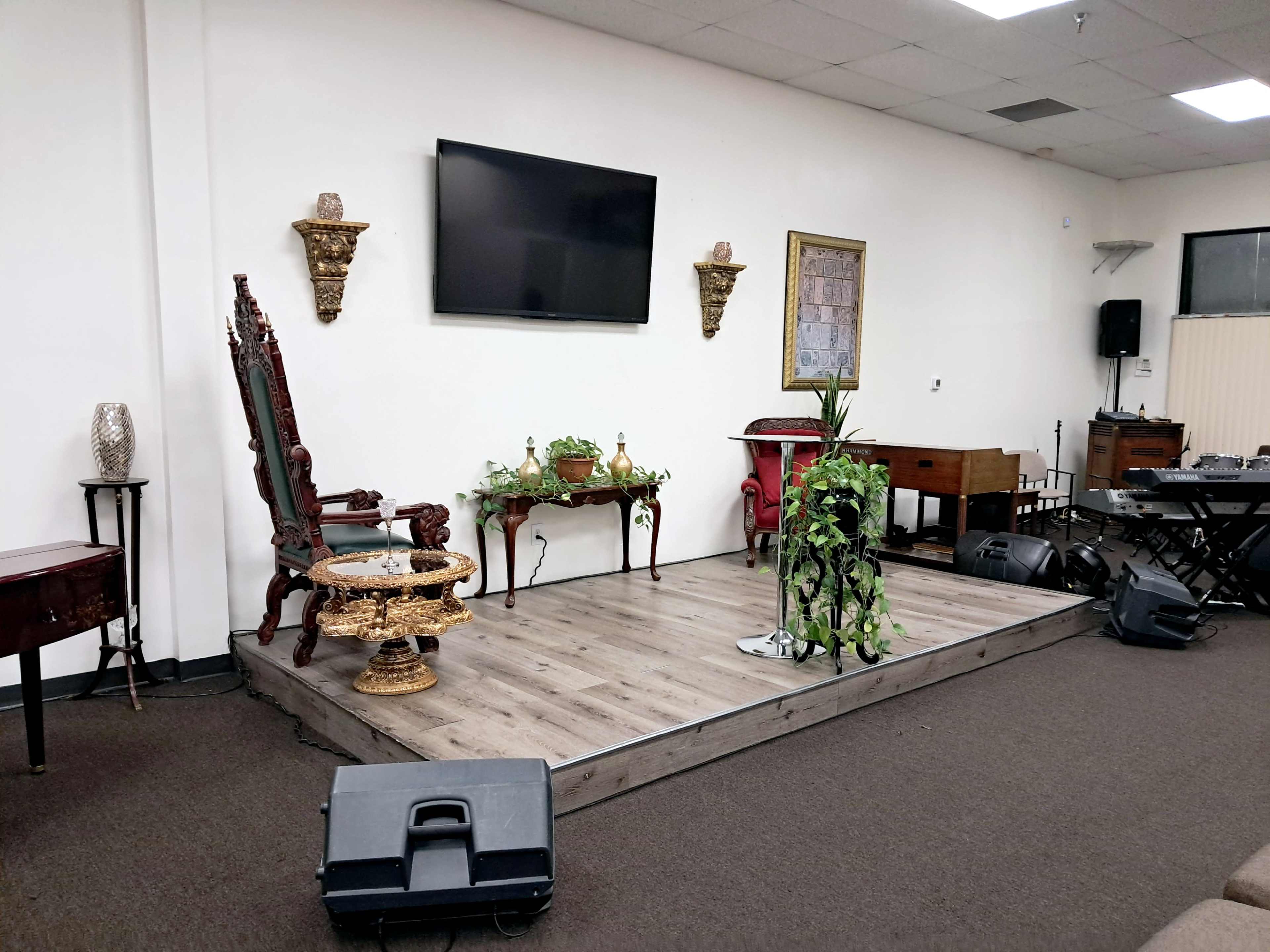 The image shows a stage area with a wooden floor, featuring a decorative chair, a small table with a plant, a television mounted on the wall, and various musical equipment in the background.