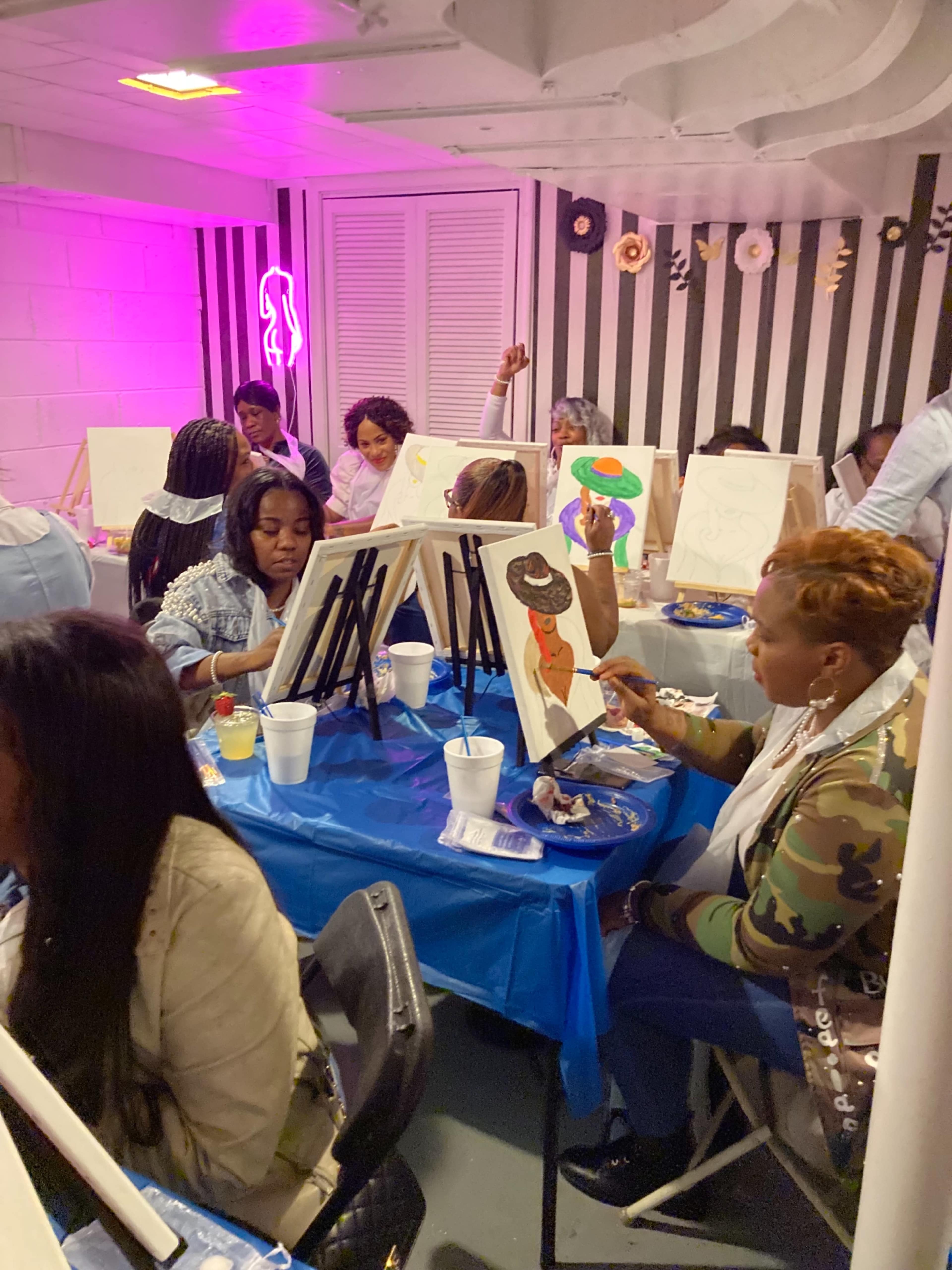 A group of people sits at tables covered with blue cloth, each painting on canvases in a spacious, brightly lit room with striped walls and decorative accents.