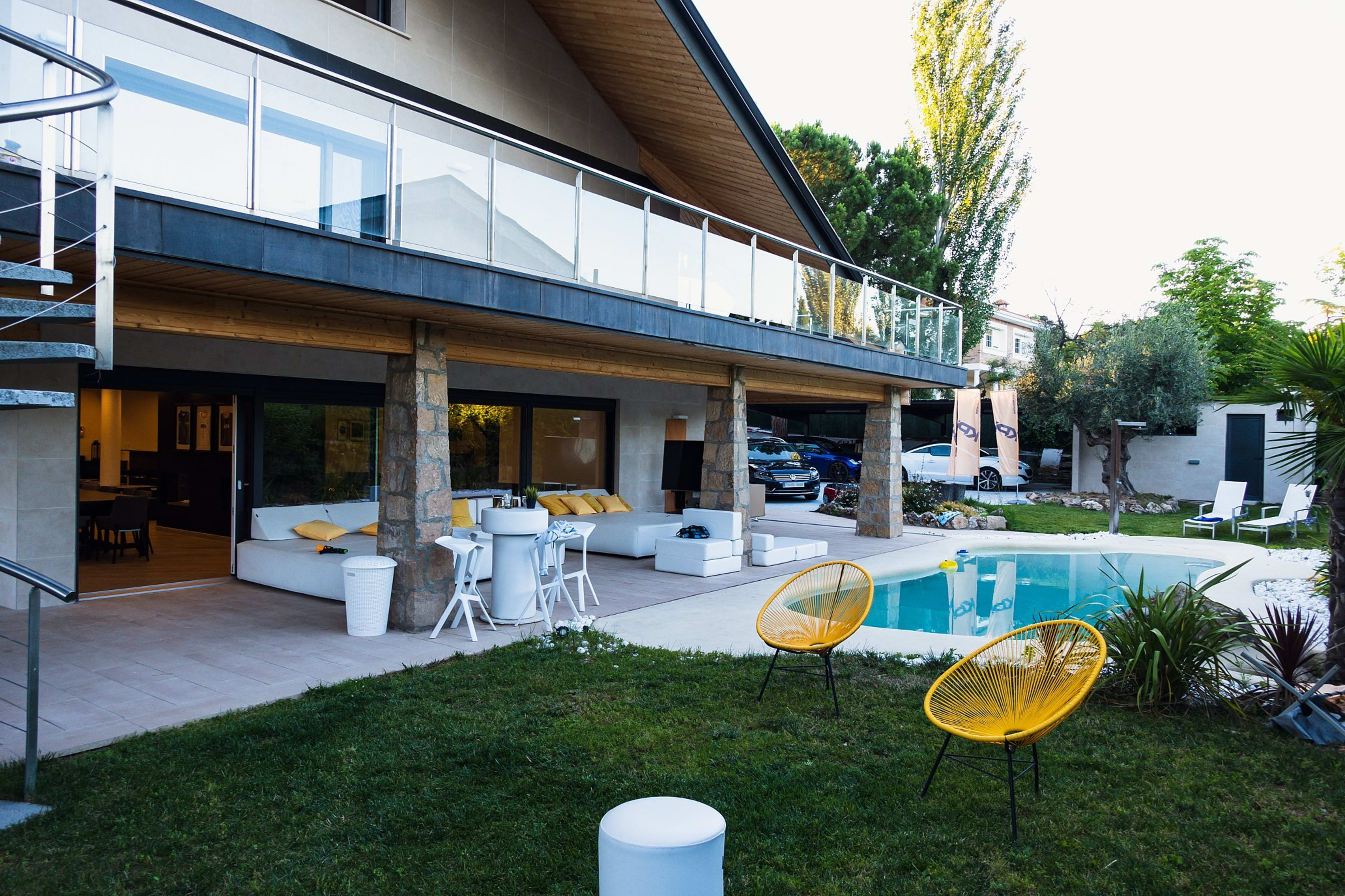 The image shows a modern house with a balcony overlooking a swimming pool and a well-kept lawn, featuring outdoor seating and landscaping.